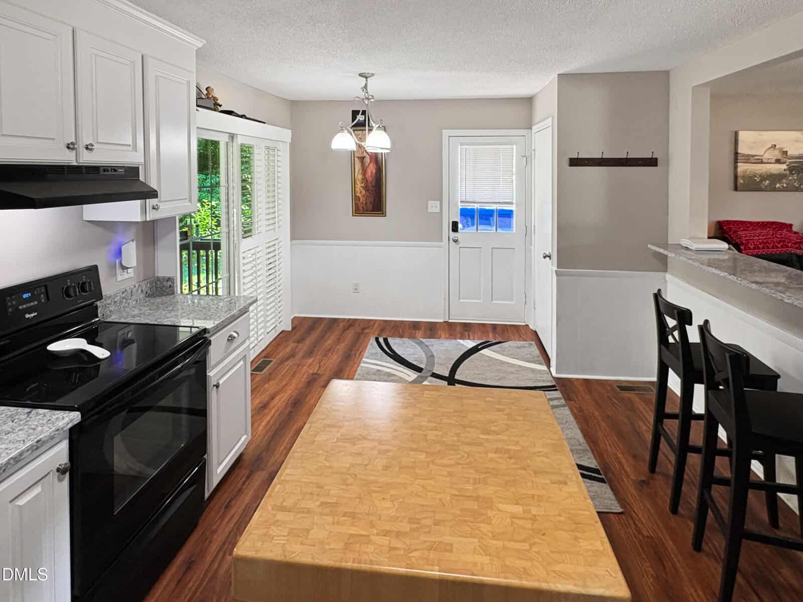 A bright kitchen and dining area with white cabinets, black appliances, wood floors, a small dining table, bar stools by a counter, and a door leading outside. Sunlight streams in through large glass doors.