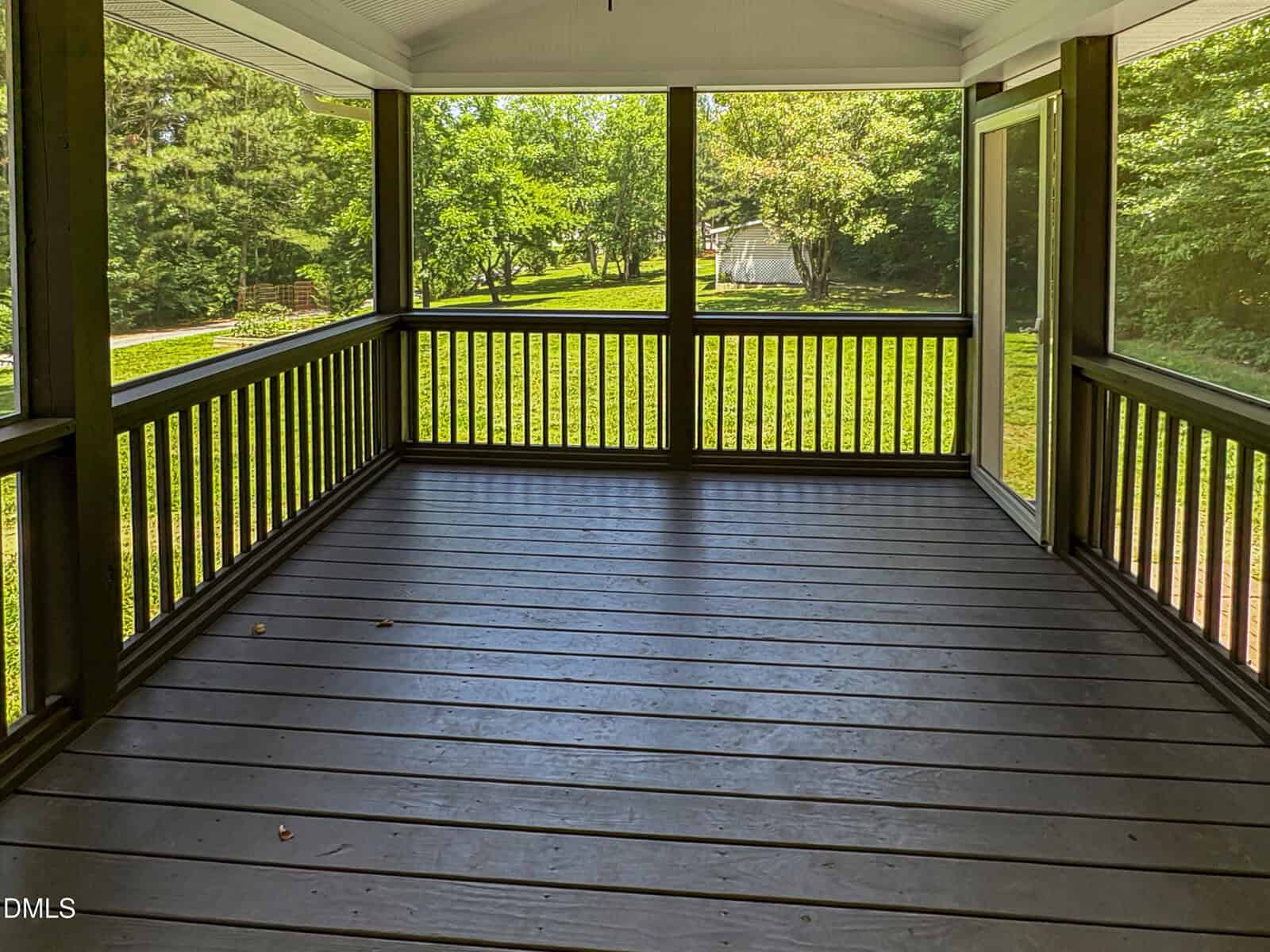 A screened-in porch with dark wooden floors and railings overlooks a sunny, green backyard with trees and a paved driveway in the distance.