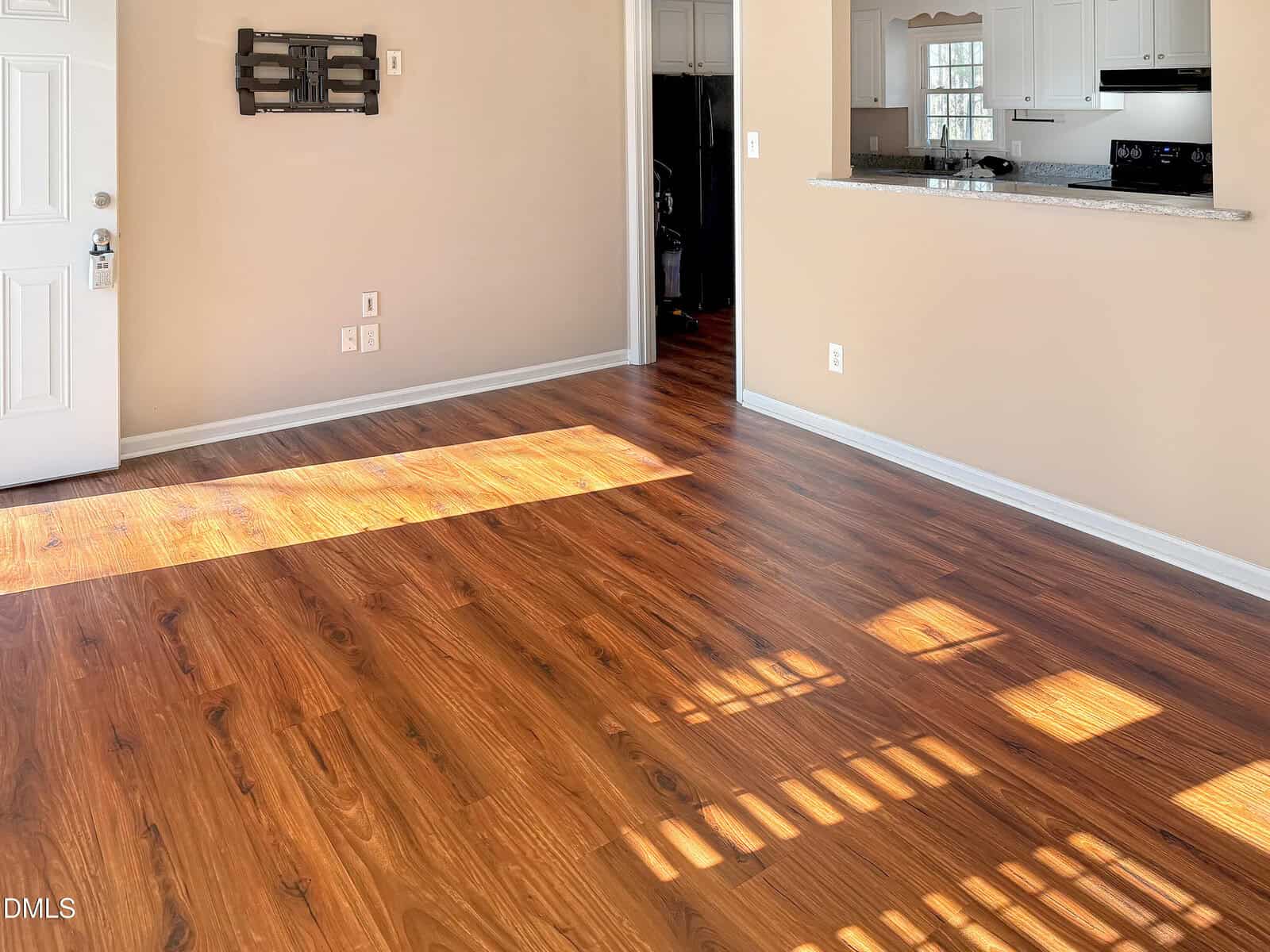 Sunlight streams onto the wood flooring of an empty living room with beige walls. A wall-mounted TV bracket is visible, and the open layout reveals part of a white kitchen with a granite countertop.