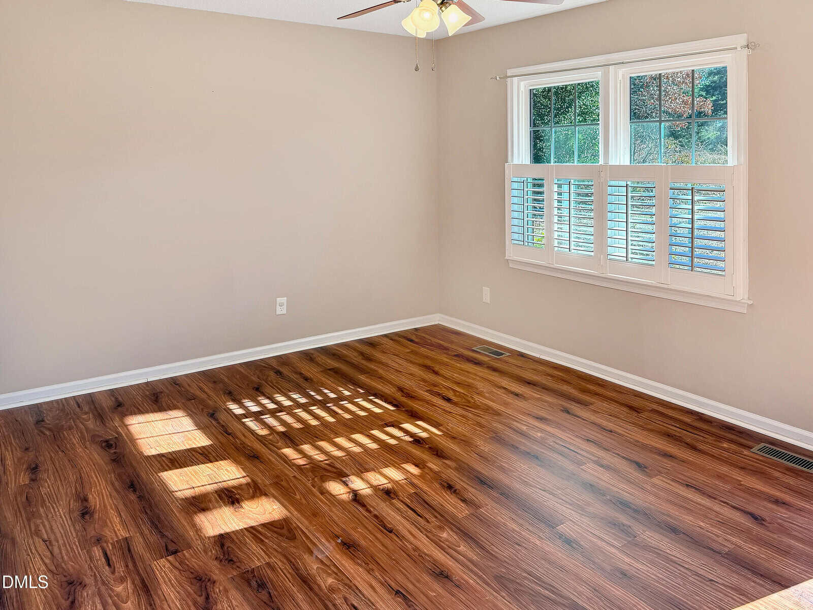 Empty room with light beige walls, a window with white shutters letting sunlight onto brown wood flooring, and a ceiling fan partially visible above.