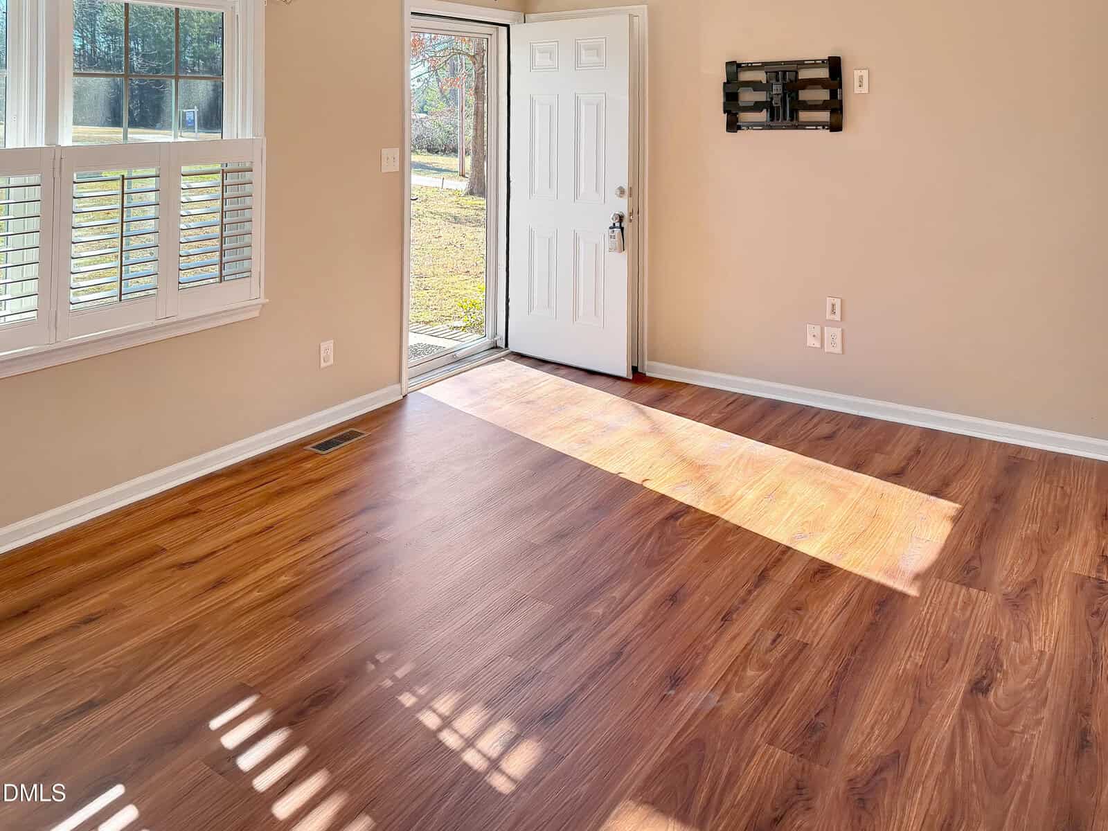 A bright, empty room with wooden floors, beige walls, a white door open to the outside, sunlight streaming in, and a window with white shutters. A TV wall mount is installed on the right wall.