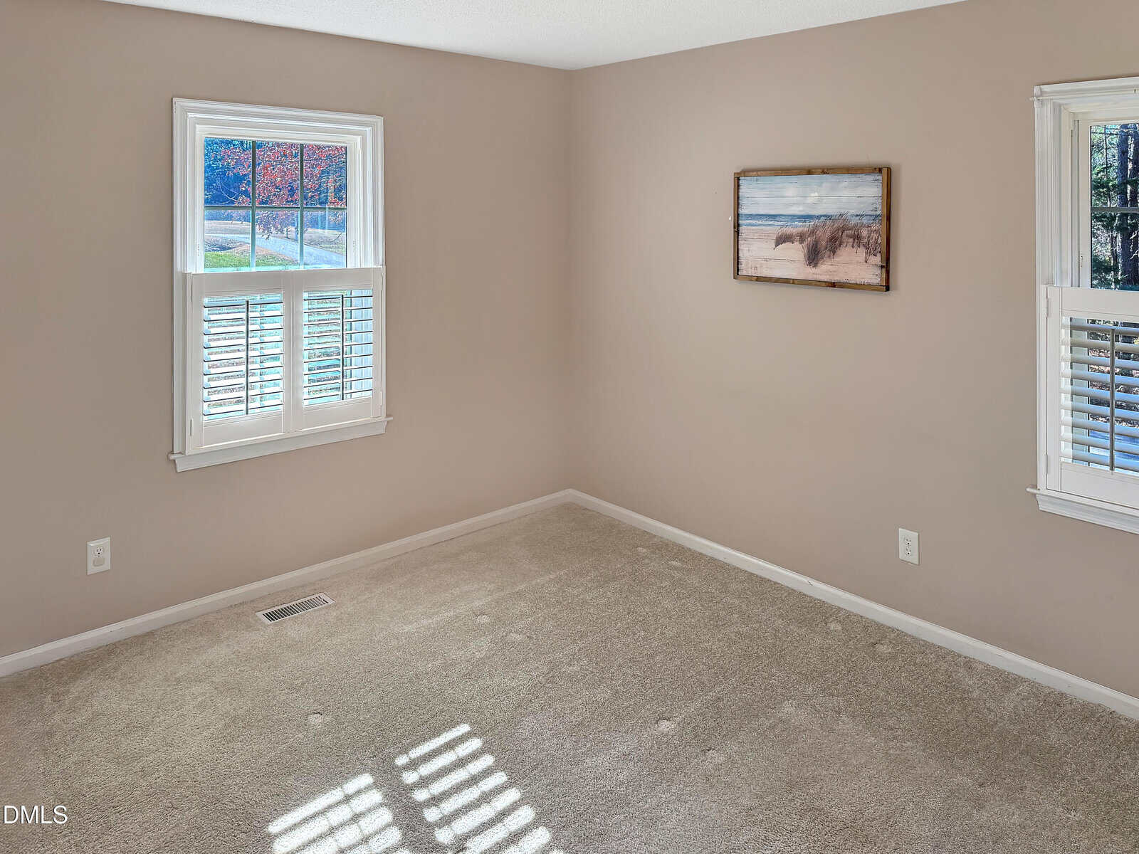 Empty room with beige carpet, light brown walls, two windows with white shutters, and a framed landscape picture hanging on the wall. Sunlight casts shadows on the carpet.