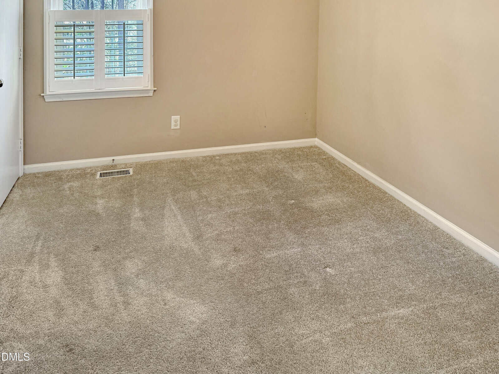 Empty beige-carpeted room with tan walls, white baseboards, a small window with white shutters, and a closed white door. There is one electrical outlet on the wall and a floor vent near the window.