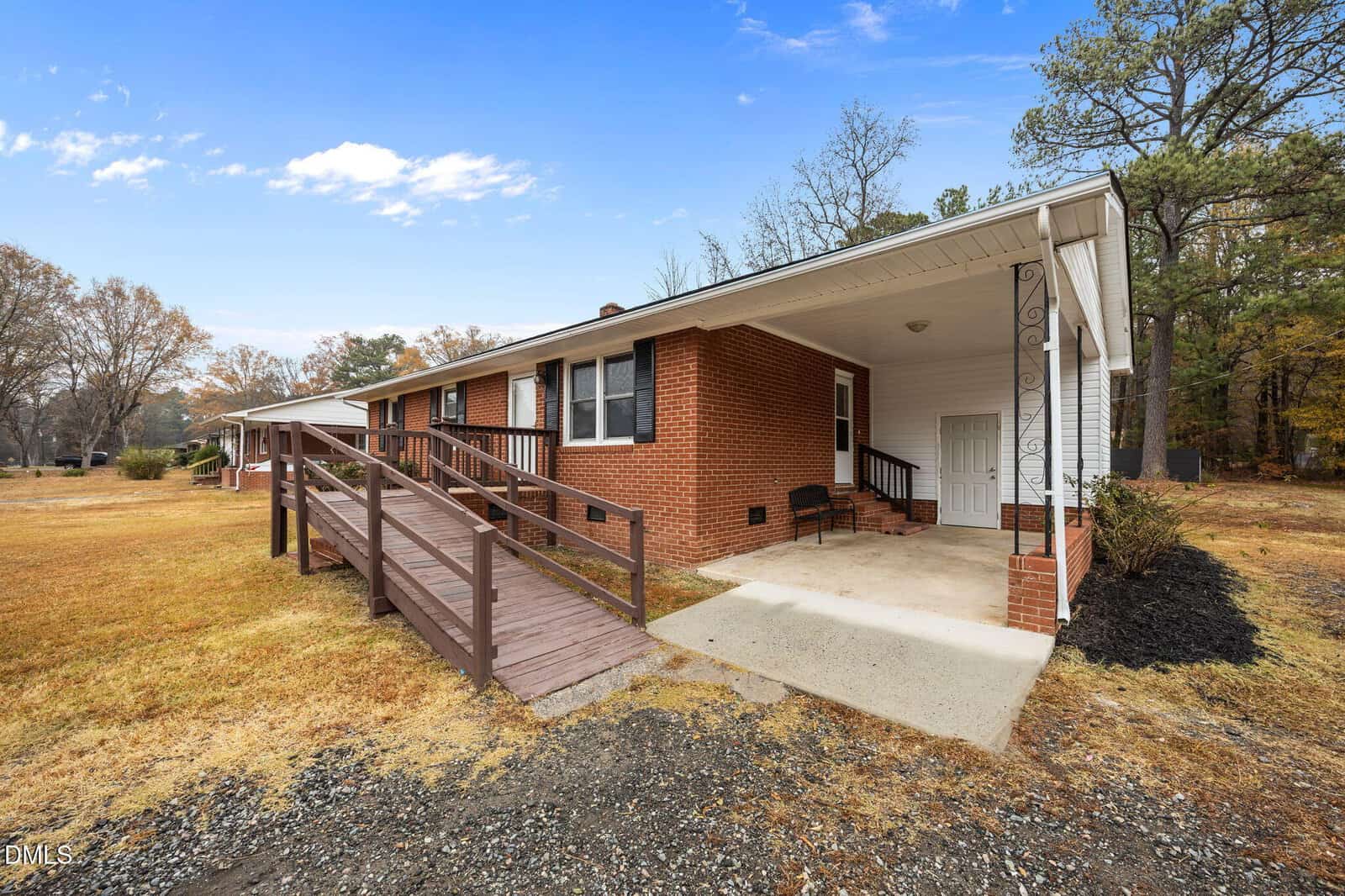 A single-story brick house with white trim, a covered front porch, and a wooden wheelchair ramp leading to the entrance. The yard is grassy with trees in the background under a blue sky.
