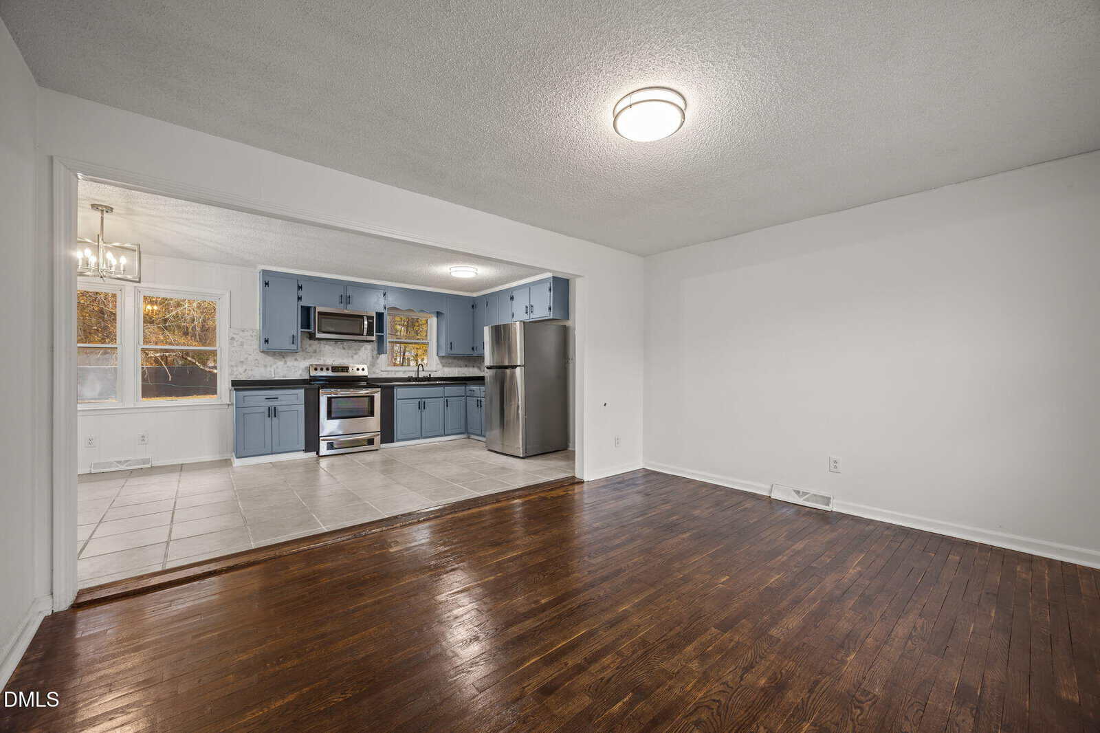 A spacious room with dark wood floors opens into a kitchen featuring blue cabinets, stainless steel appliances, tile flooring, and a window by the dining area. The walls are white, and ceiling lights are on in both areas.