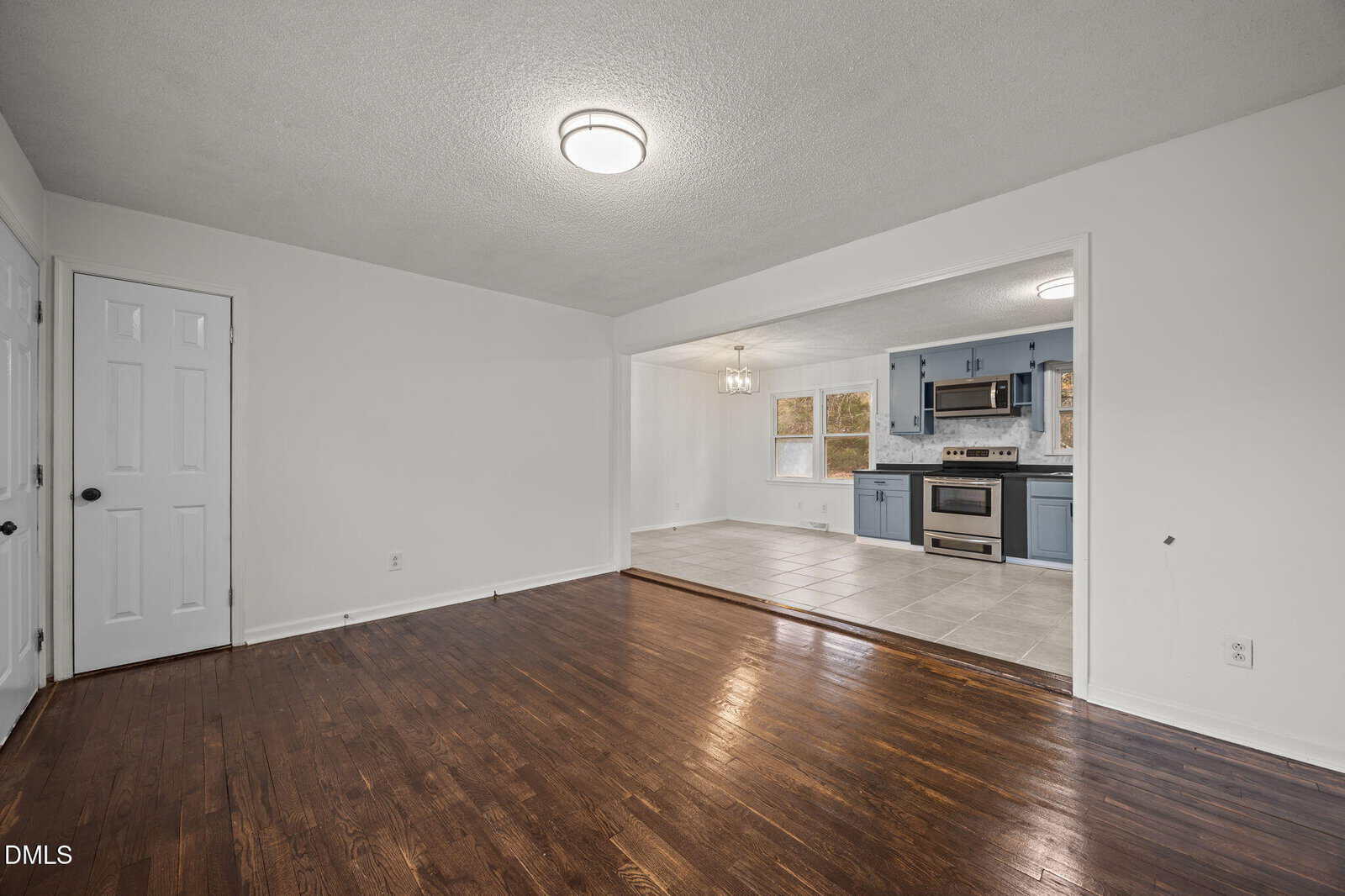 A view of an empty room with dark wood floors leading into a kitchen area. The kitchen features gray cabinets, stainless steel appliances, tile flooring, and a hanging light fixture over a dining space. White walls throughout.