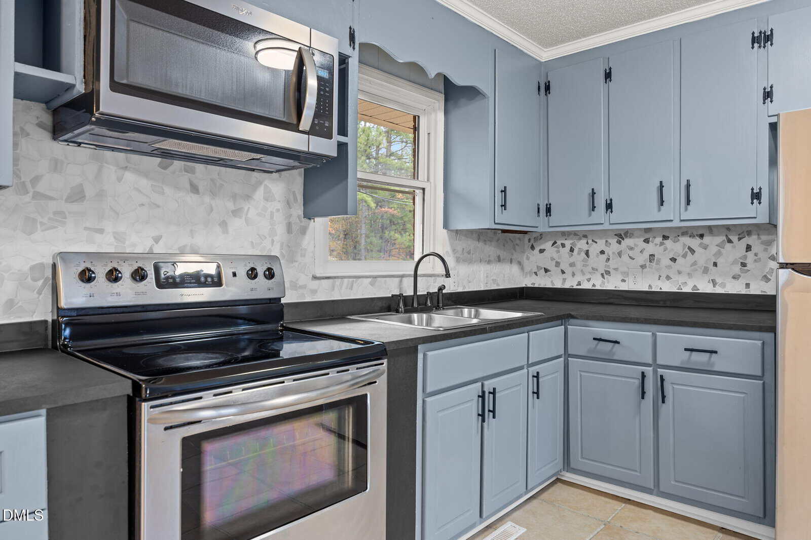 Modern kitchen with blue cabinets, black countertops, a stainless steel stove, microwave, and a double sink beneath a window. Mosaic tile backsplash and neutral tiled floor are also visible.