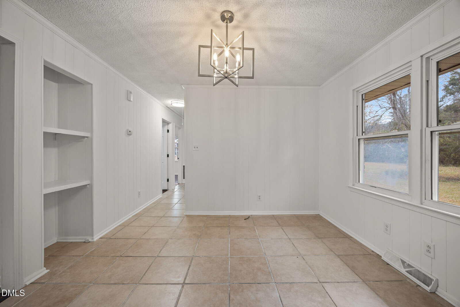 A small, empty room with beige tile flooring, white paneled walls, built-in shelves, a modern light fixture, and a window overlooking a grassy yard.