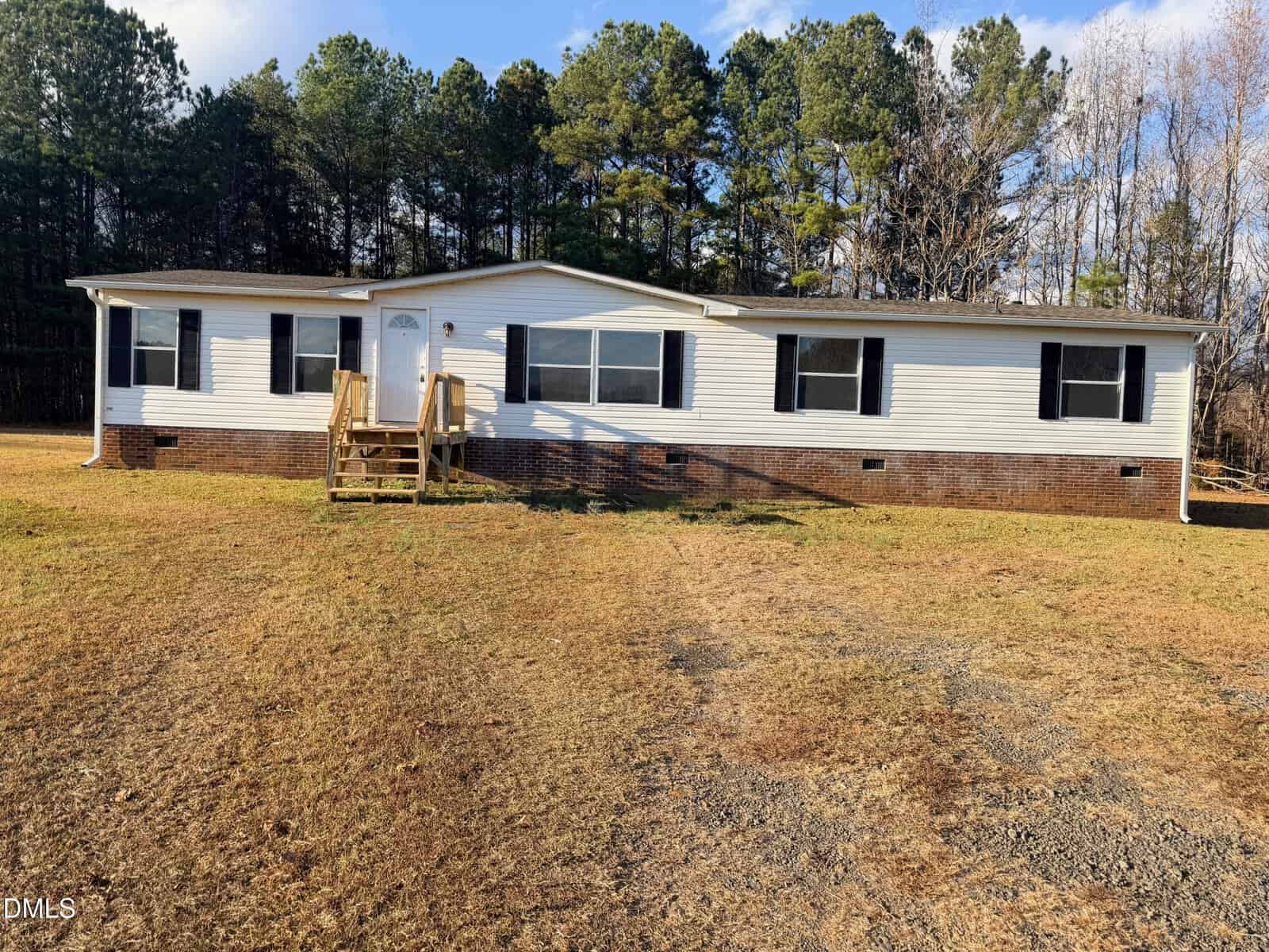 A single-story manufactured home at 1185 Diamond Hills Road, Stem, with white siding, black shutters, and a brick foundation. Wooden steps lead to the front door. The house sits on a grassy lot with trees under a partly cloudy sky.