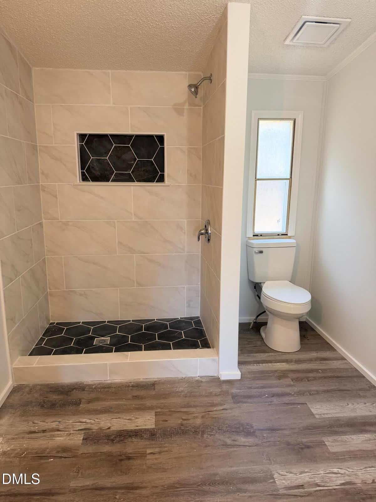 A bathroom with a walk-in shower featuring large beige tiles, a black hexagonal tile floor and niche, a white toilet next to a window, and wood-look vinyl flooring.