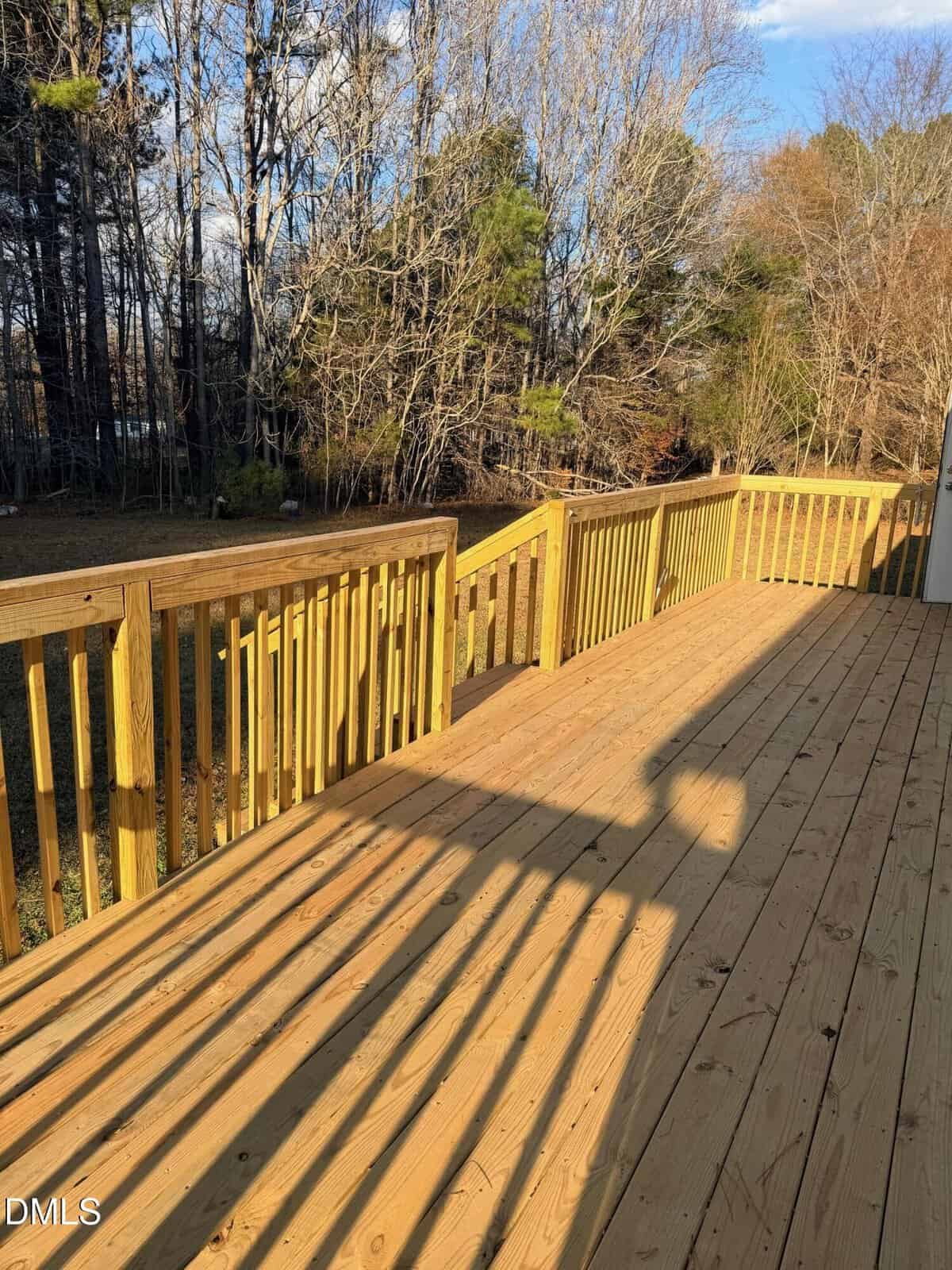 A wooden deck with a yellow railing overlooks a grassy backyard bordered by leafless trees under a partly cloudy sky. Long shadows are cast across the deck.