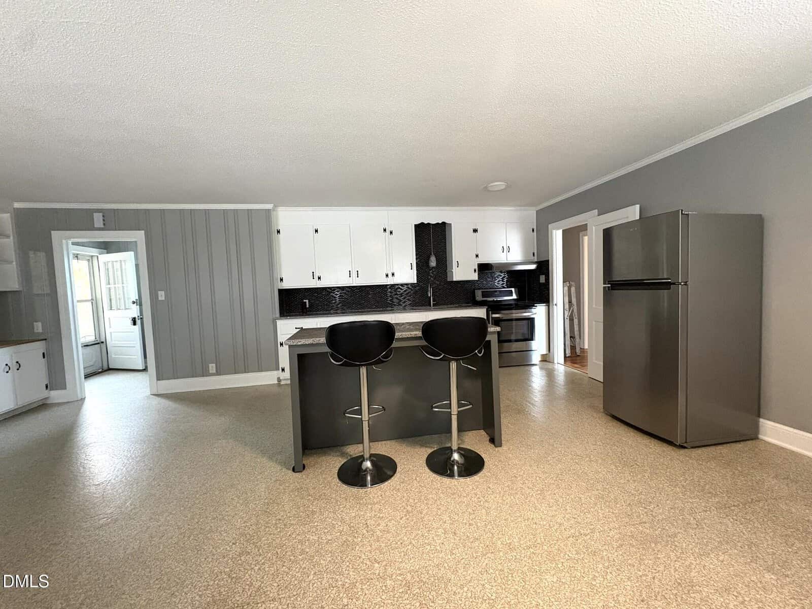 A modern kitchen with gray walls, white upper cabinets, black lower cabinets, a large island with two black barstools, stainless steel appliances, and beige flooring. A door leads to another room on the left.