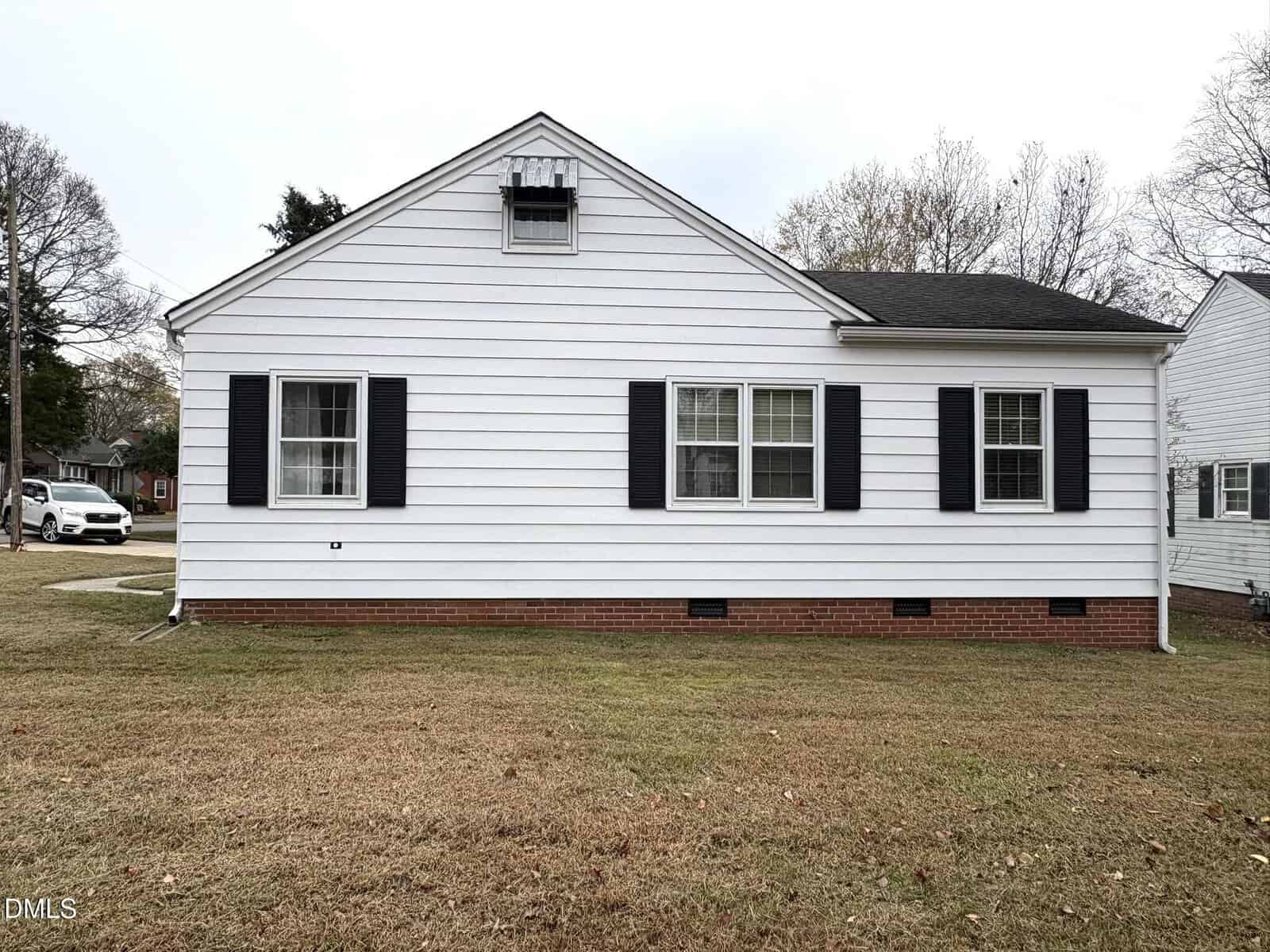 A single-story white house with horizontal siding, black shutters, and a brick foundation, situated on a brown lawn. A white car is parked on the street to the left, and trees are visible in the background.
