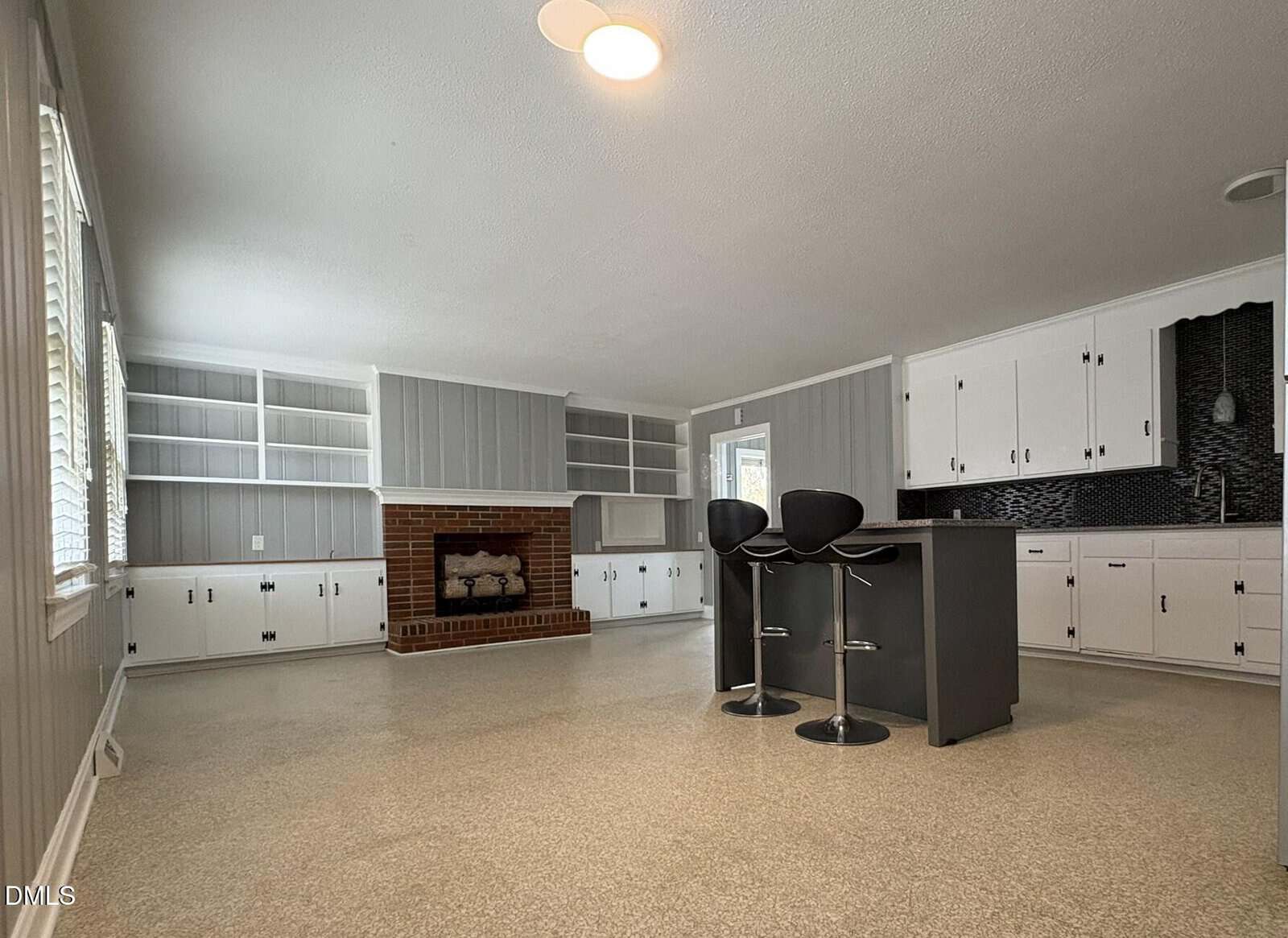 Spacious kitchen and living area with gray walls, white cabinets, a brick fireplace, built-in shelves, an island with two black barstools, and a window letting in natural light.