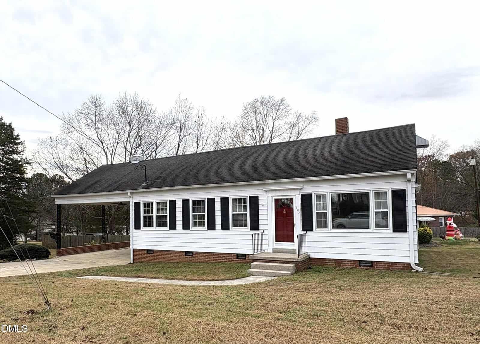 A single-story white house with black shutters and a red door, featuring a carport on the left side. The yard has patchy grass, and there are leafless trees and another house visible in the background.