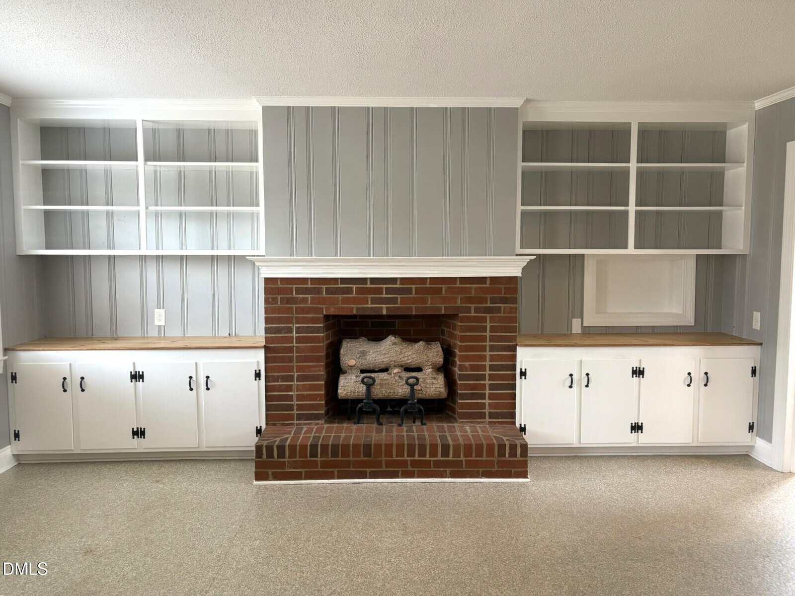 Living room with a central red brick fireplace, flanked by white built-in cabinets and open shelves. The walls are gray with vertical paneling, and the floor is a light neutral color.