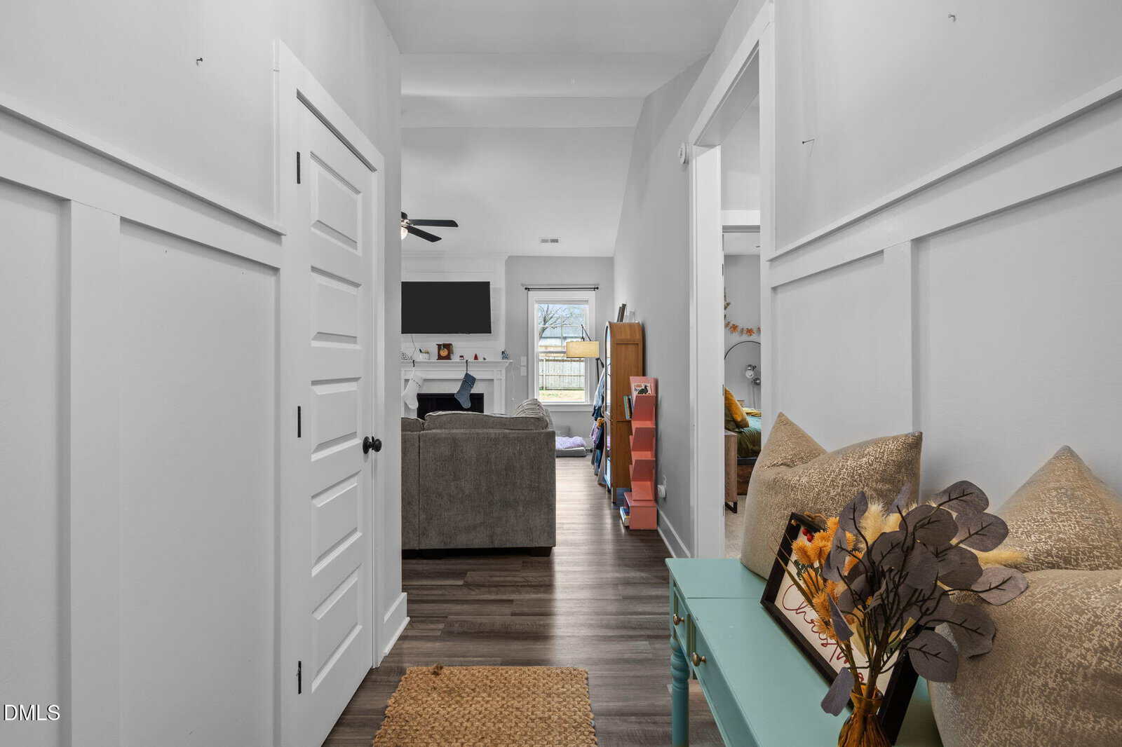 A view down a hallway with white walls and wood flooring, leading to a living room with a sofa, wall-mounted TV, and fireplace. A glass table with pillows and decorative branches is on the right.