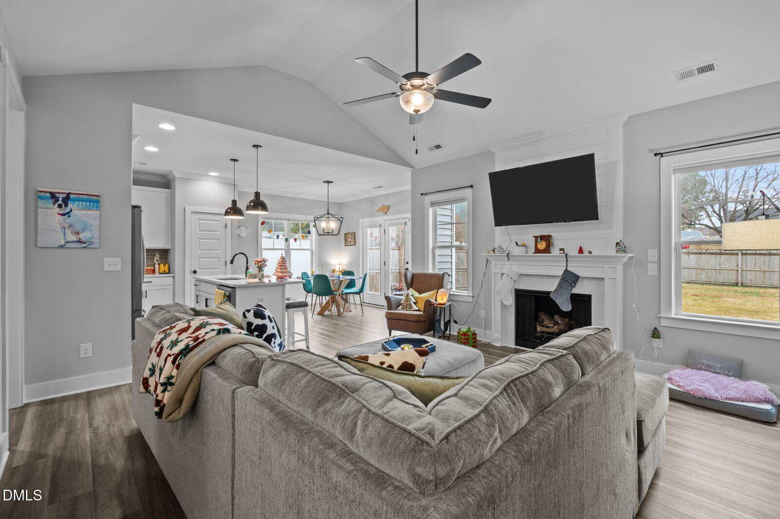 A modern living room with a large gray sectional sofa, a ceiling fan, a wall-mounted TV above a white fireplace, and adjacent open kitchen and dining areas. Large windows let in natural light.