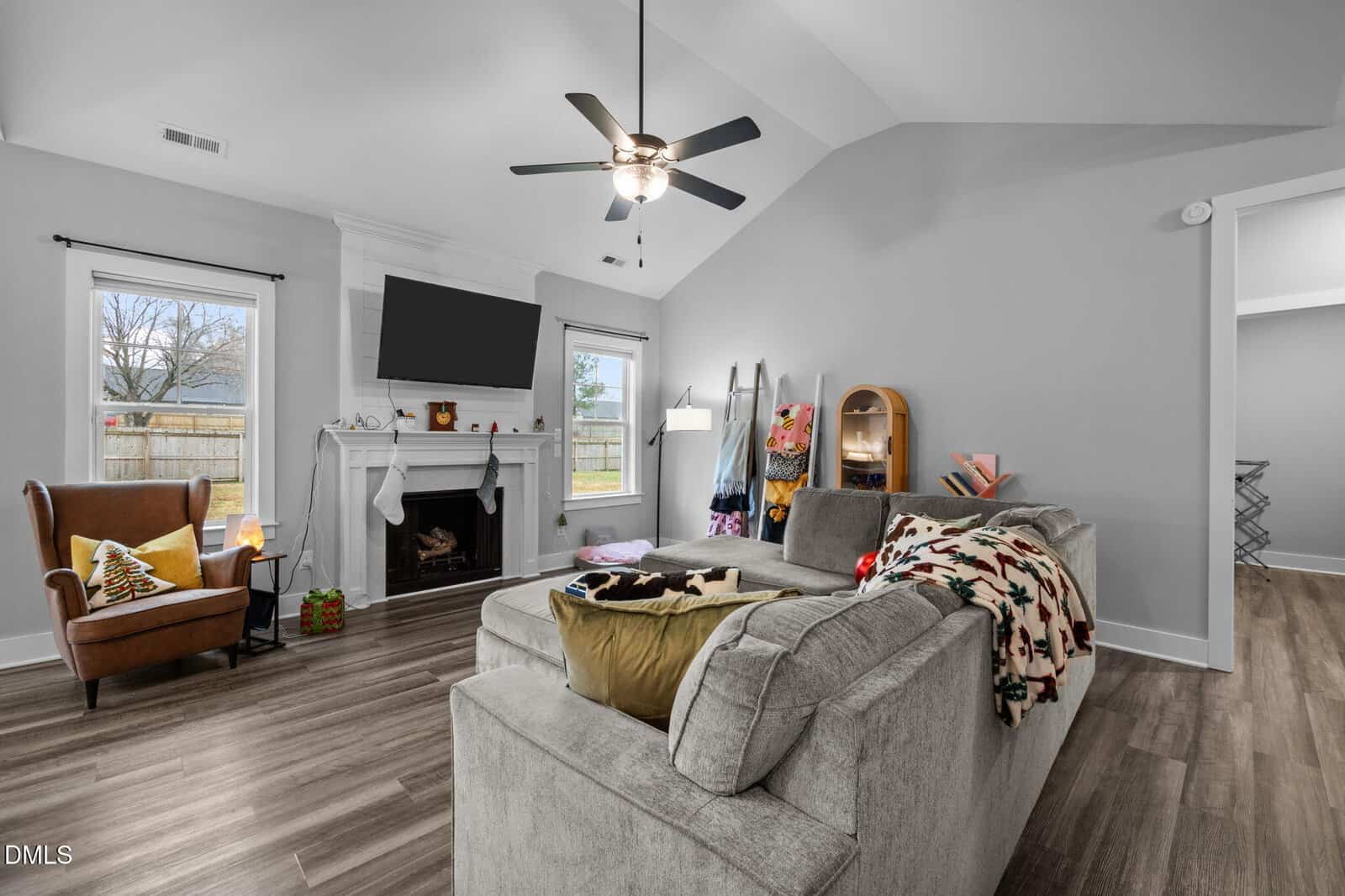 A modern living room with gray walls and a vaulted ceiling. Features a sectional sofa, armchair, fireplace with TV above, ceiling fan, and wood flooring. Windows let in natural light; various decor items are arranged throughout the space.