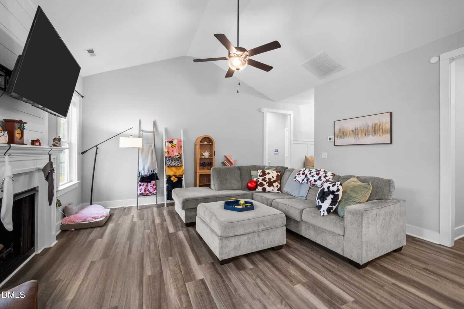A living room with gray sectional sofa, patterned pillows, a ceiling fan, mounted TV, fireplace, and wooden floors. Shelves with blankets and a pet bed are against a white wall near a window.