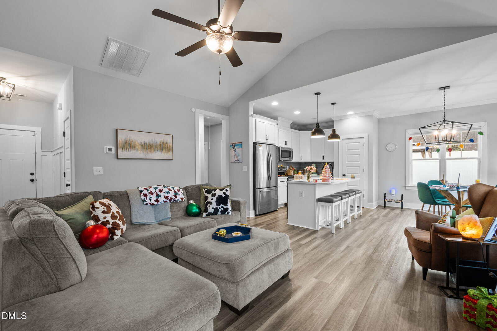 A modern living room with a gray sectional sofa, holiday decorations, open kitchen with an island and barstools, stainless steel appliances, and pendant lights, featuring wood flooring and neutral walls.