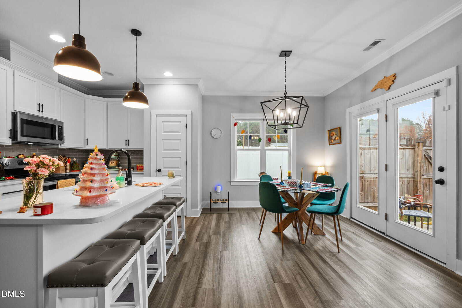 A modern kitchen and dining area with white cabinets, an island with four stools, a decorated cake on the counter, a round dining table with green chairs, wood flooring, and large windows letting in natural light.