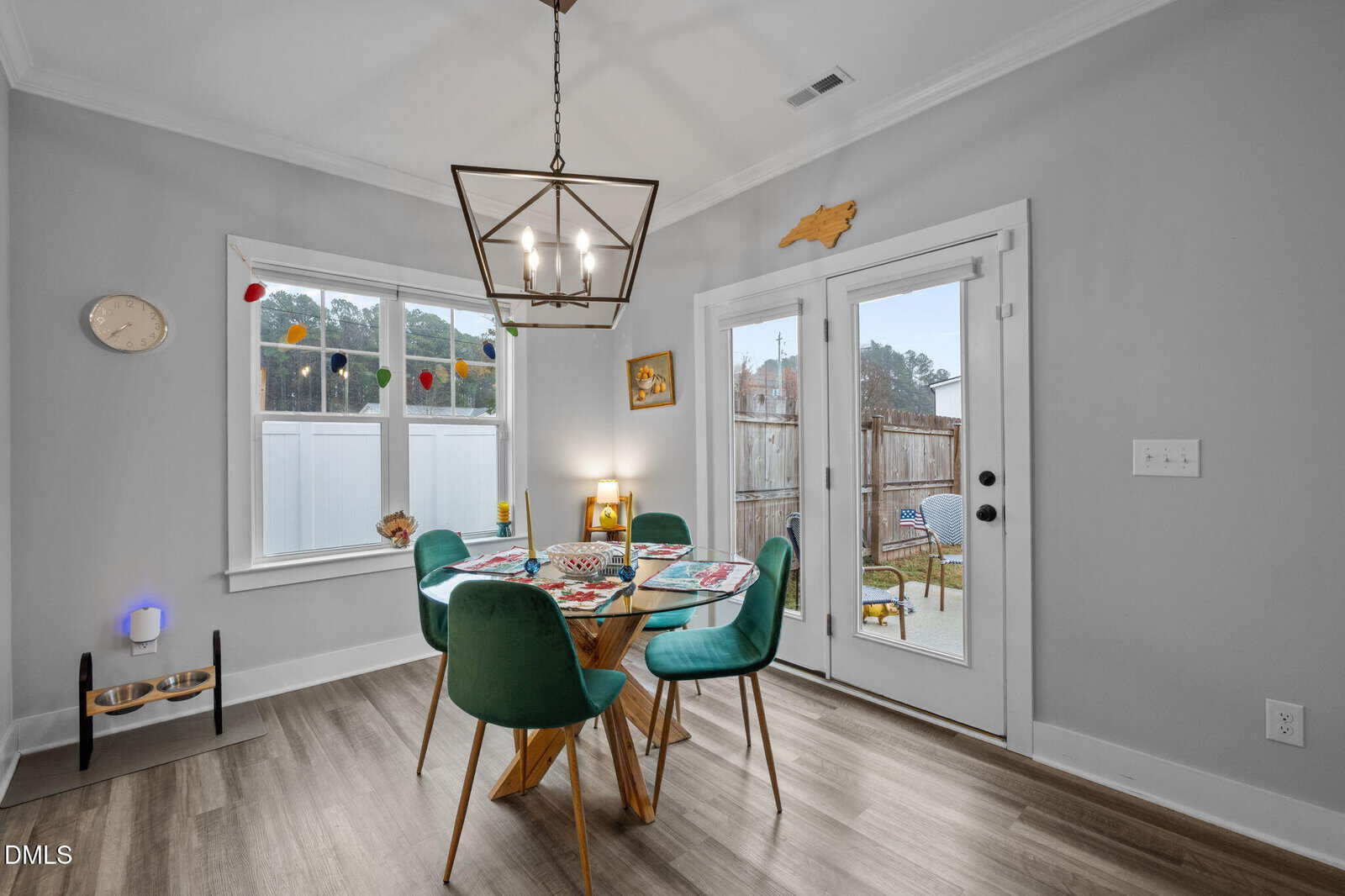 A dining area with a round wooden table, four teal chairs, placemats, and decor. A geometric light fixture hangs above. French doors lead outside, and a window with colorful pom-pom garland is on the left.