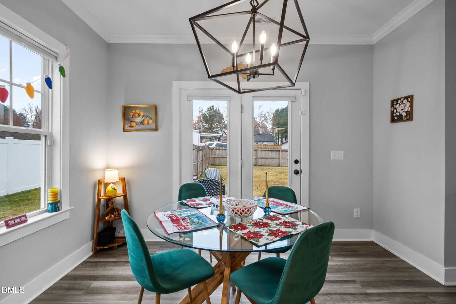 A dining area with a round glass table and four teal chairs sits near double glass doors leading outside. The room has light gray walls, wood flooring, and a geometric chandelier above the table.