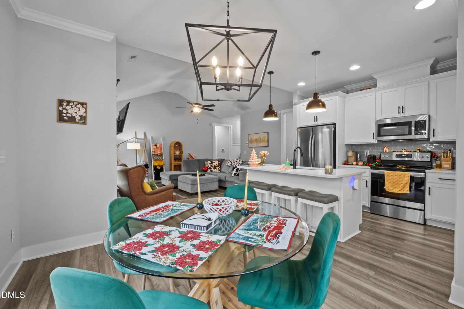 A modern open-concept kitchen and living area with a glass dining table, teal chairs, floral placemats, a white island with stools, stainless steel appliances, and neutral-toned furnishings and decor.