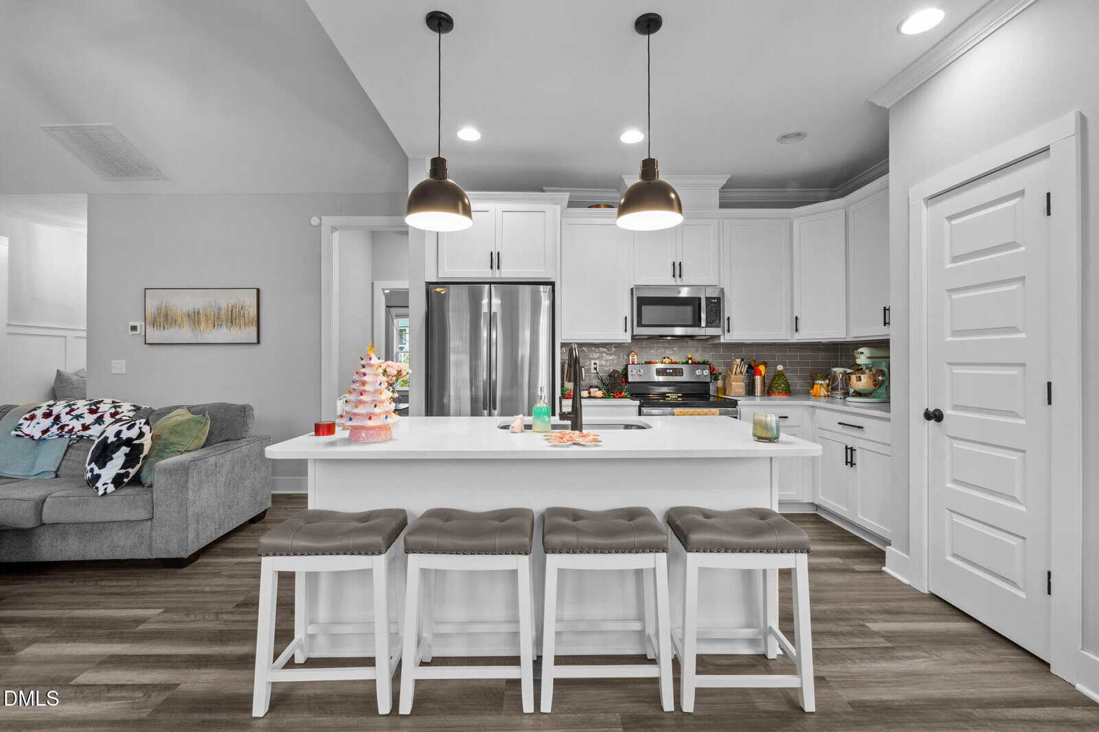 Modern kitchen with white cabinets, stainless steel appliances, a large island with four gray cushioned stools, pendant lights, and festive decor. A gray sofa with a colorful blanket sits to the left.