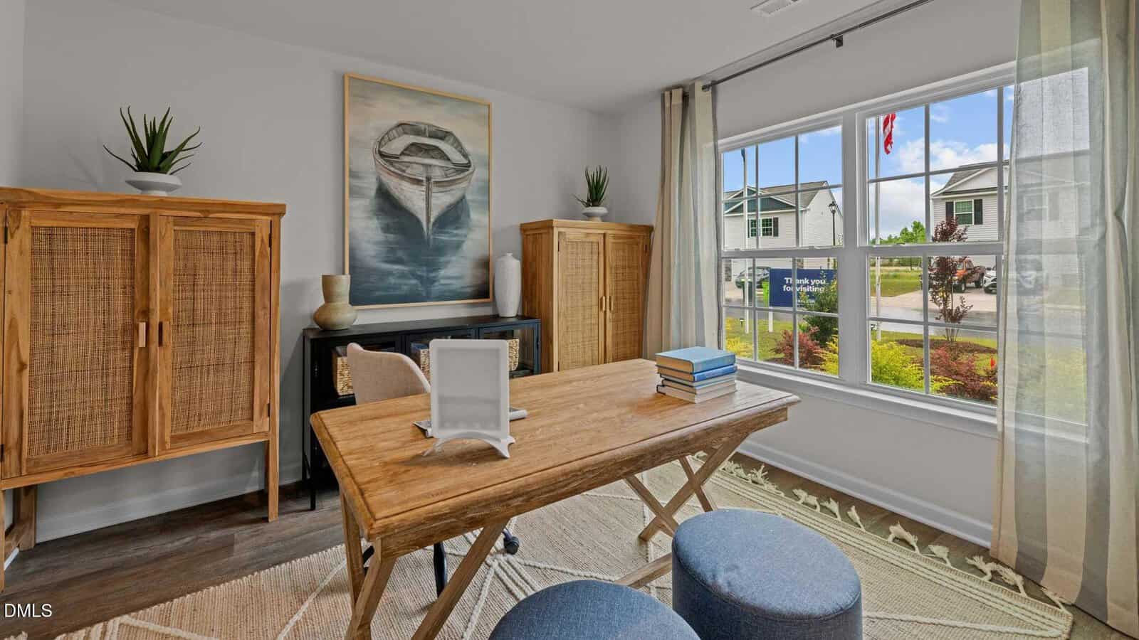 A bright home office with a wooden desk, two blue ottomans, two wooden cabinets, a painting of a boat on the wall, books on the desk, and a large window overlooking a suburban street.