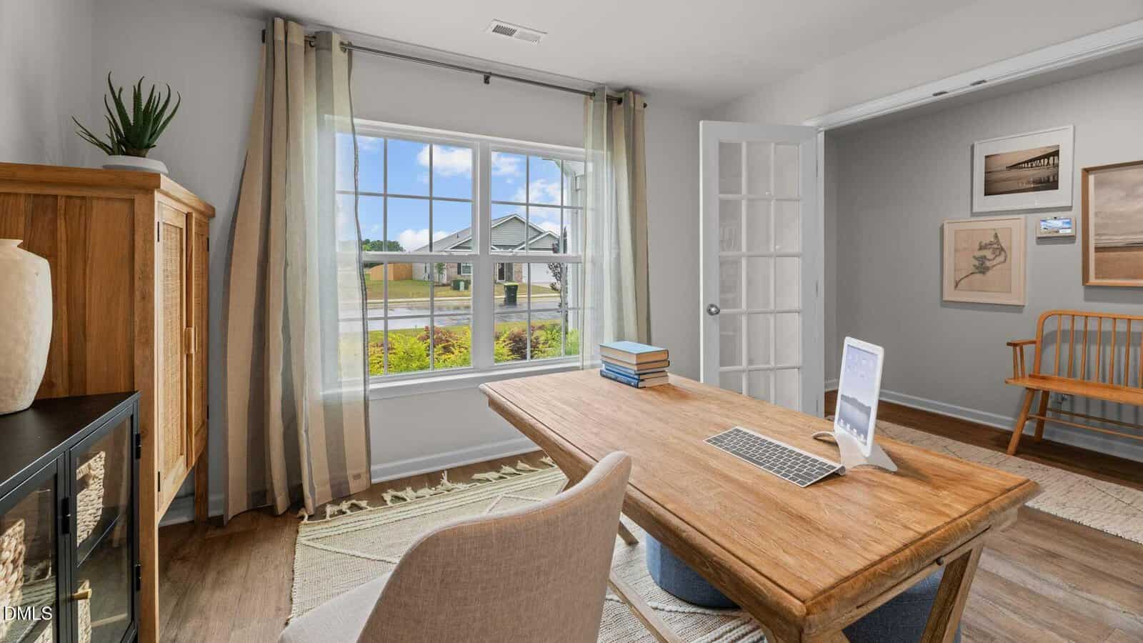 A home office with a wooden desk, chair, laptop, and stacked books. Large window with curtains lets in natural light. There is a cabinet, plant, wall art, and a wooden bench against a light-colored wall.