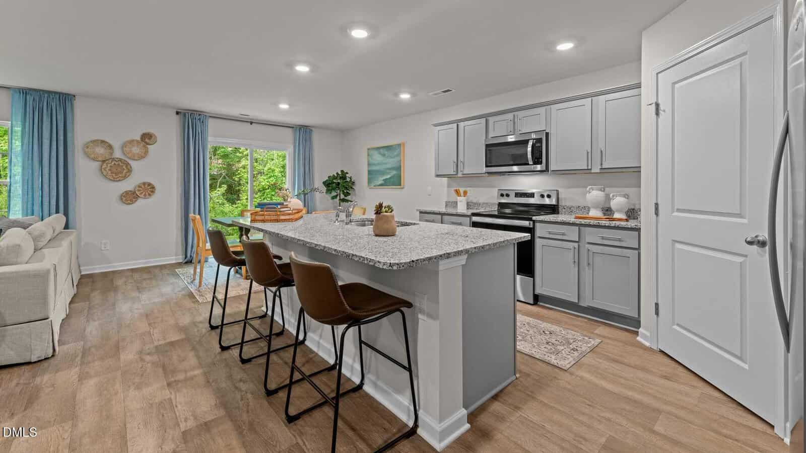 A modern kitchen with light gray cabinets, stainless steel appliances, and a central island with three brown barstools. The room features wood flooring and a living area with a beige sofa and blue curtains in the background.