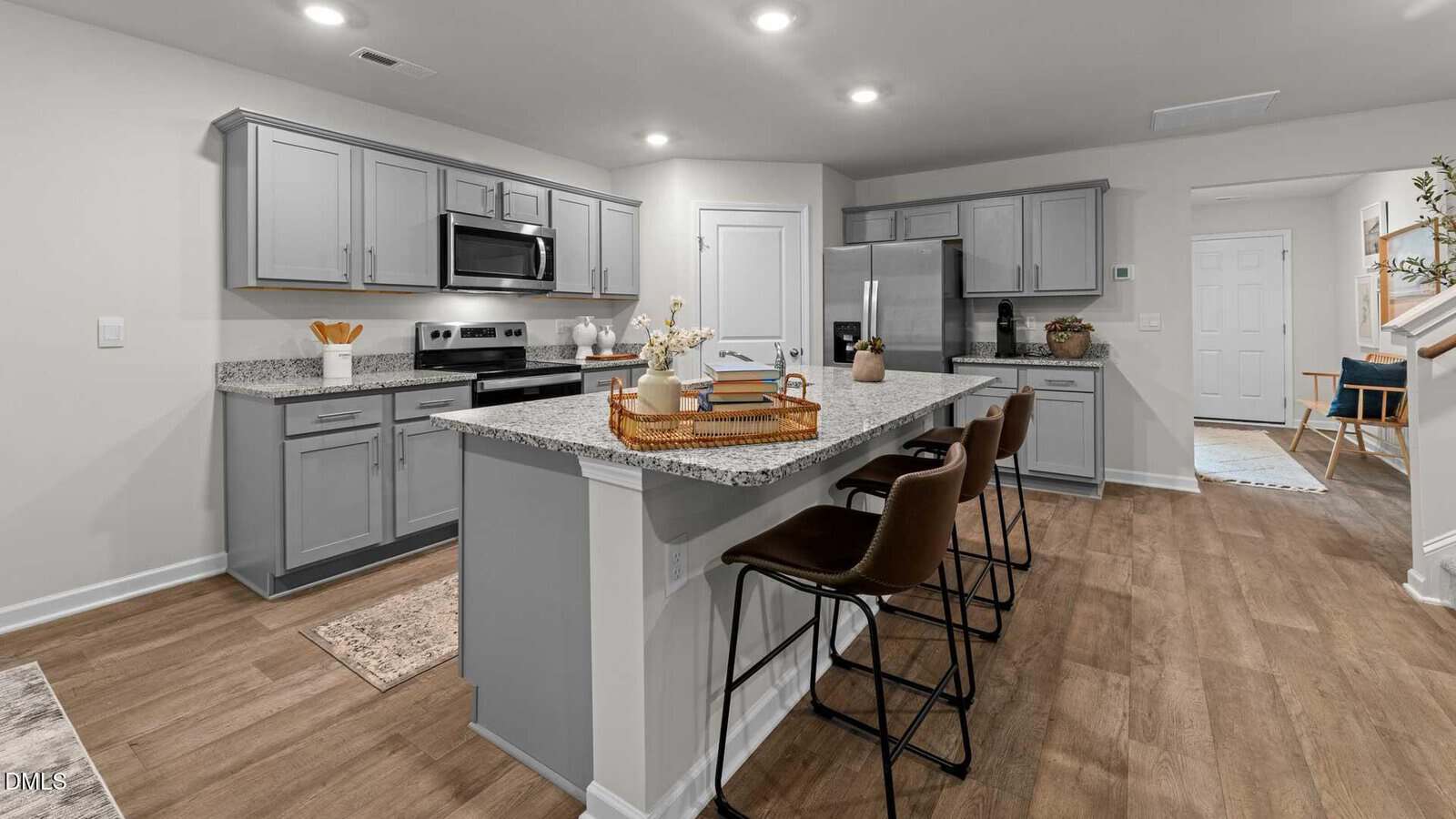 A modern kitchen with gray cabinets, stainless steel appliances, a granite island with a wicker tray, and three brown chairs. The floor is wood, and the walls are light-colored with recessed lighting overhead.