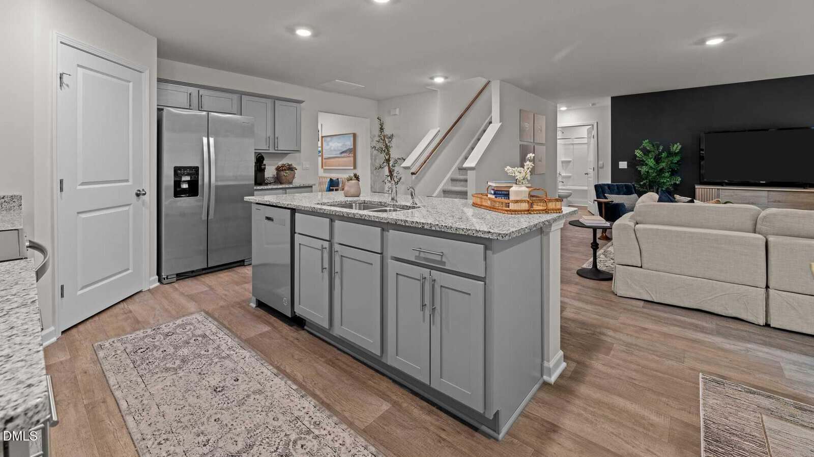 Modern kitchen with gray cabinets, granite countertops, stainless steel refrigerator, island with sink, and open layout leading to a living area with beige sofas, stairs, and a black accent wall with a TV.