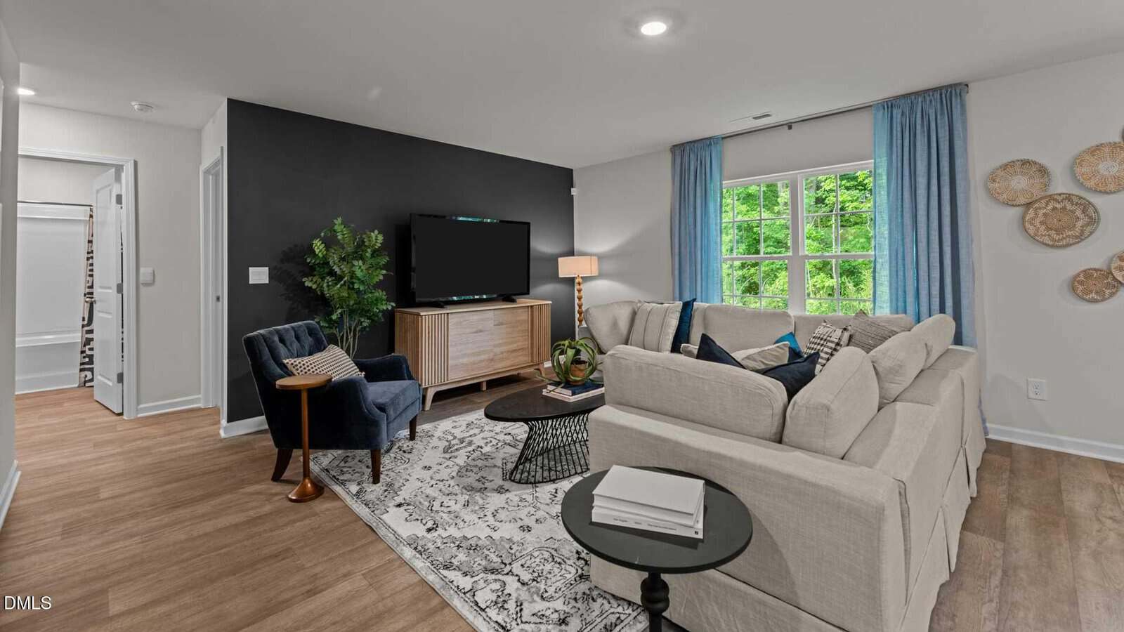 A modern living room with a beige sectional sofa, blue accent chair, wooden TV stand, indoor plant, and a patterned rug. The room features blue curtains, wall decor, wood flooring, and a window showing green trees outside.