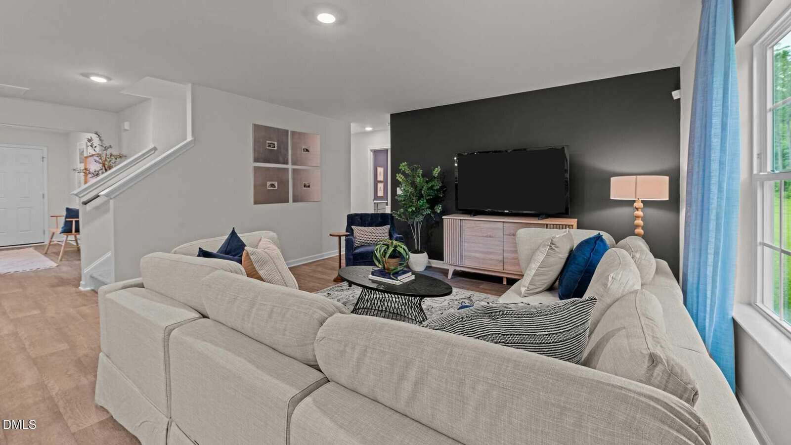 A modern living room with a beige sectional sofa, navy and striped throw pillows, a black accent wall, a flat-screen TV on a wooden console, a lamp, wall art, and a window with blue curtains.
