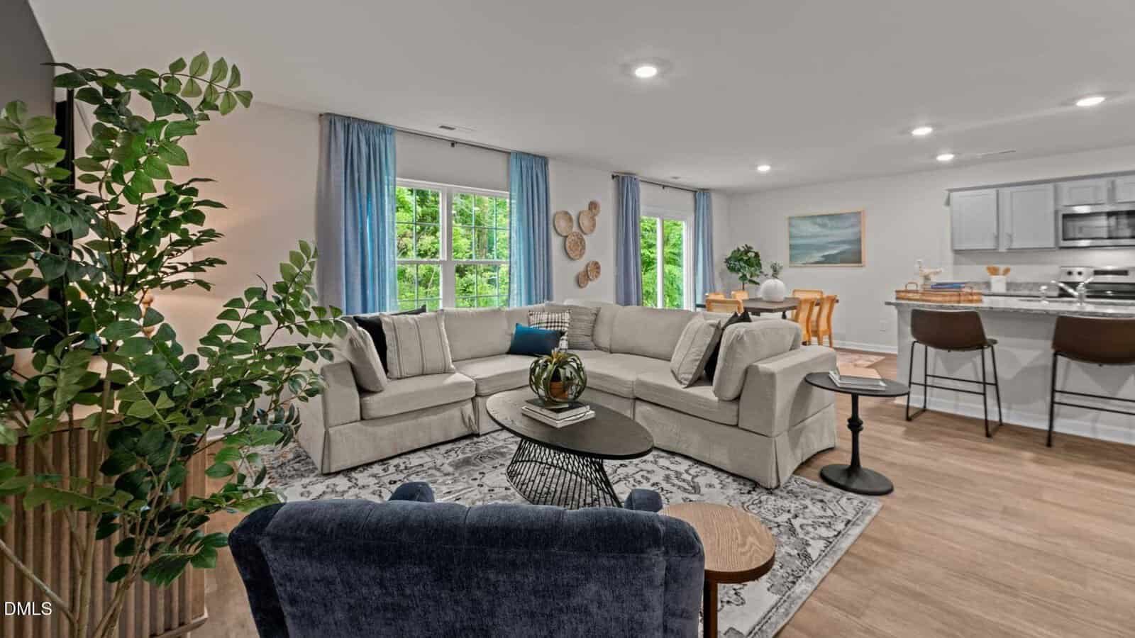 A modern living room with a large beige sectional sofa, blue armchair, coffee table, and potted plant. Behind are windows with blue curtains, a dining table, and an open kitchen with bar stools and light wood flooring.