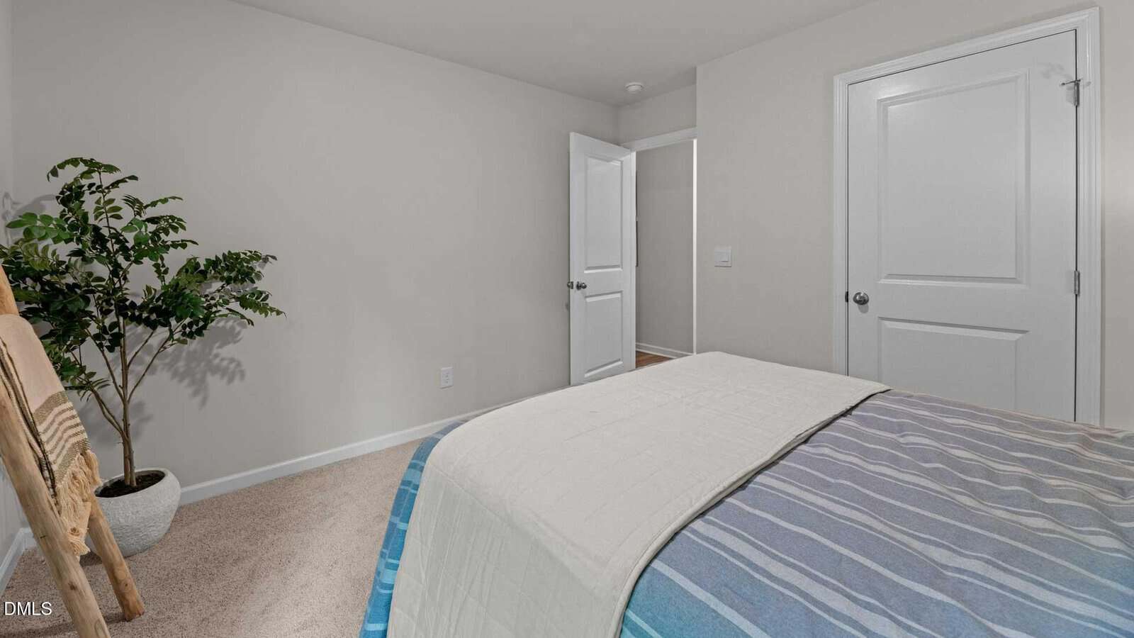 A neatly made bed with striped bedding in a simple bedroom with beige walls, two closed white doors, a green potted plant, and a wooden blanket ladder in the corner. Carpeted floor and minimal decor are visible.