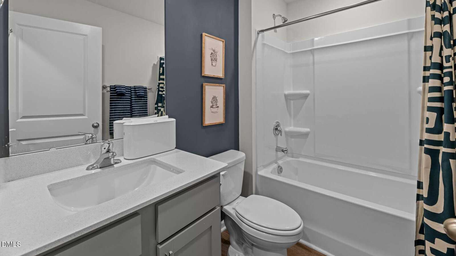 Modern bathroom with a white sink and cabinet, toilet, bathtub with shower, and geometric shower curtain. Two framed pictures hang on a blue accent wall above the toilet. Towels are stacked near the door.