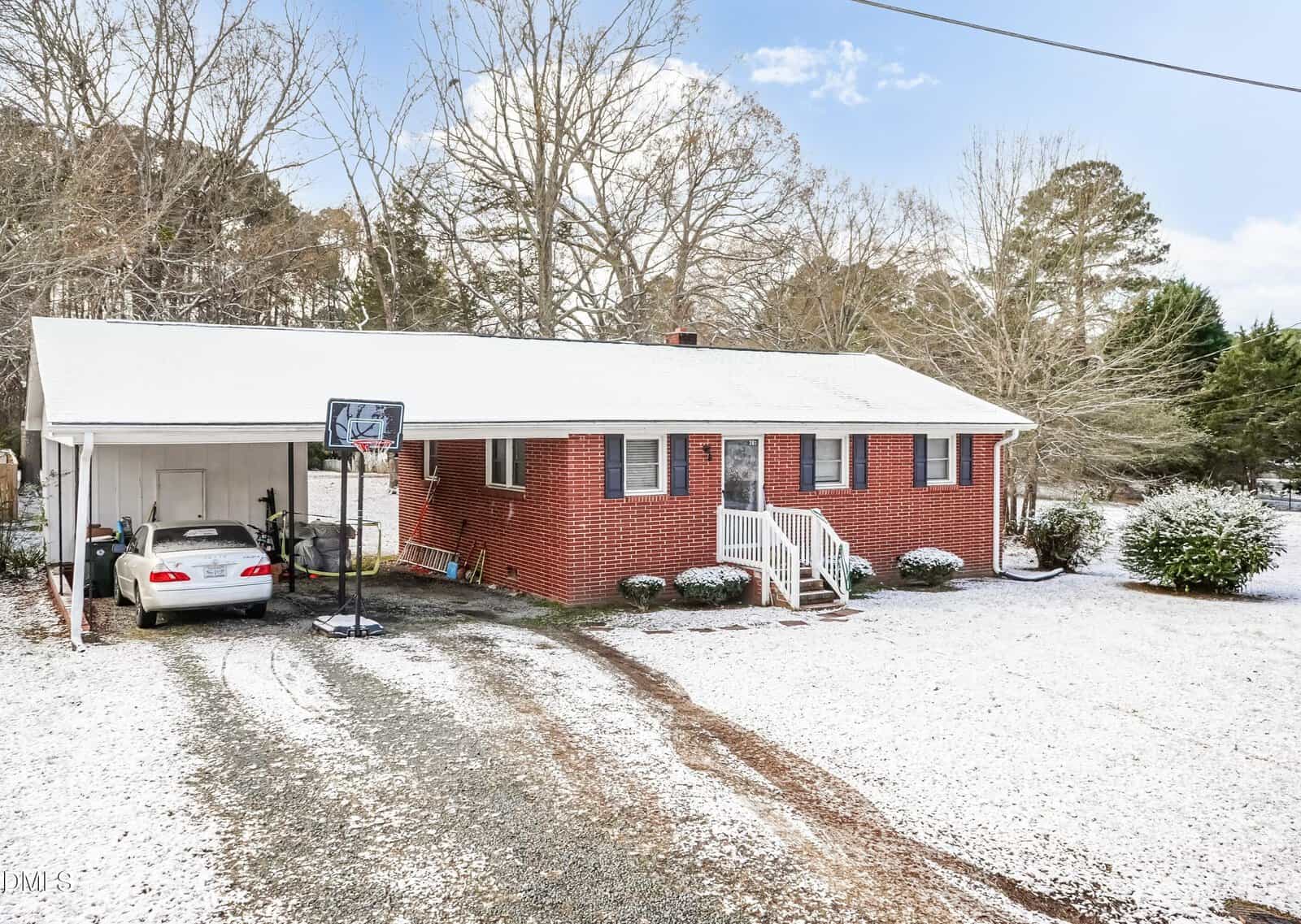 A red brick single-story house at 207 Rogers Avenue in Creedmoor features a white front porch overlooking a snowy yard. A car rests under the left carport, while a basketball hoop stands by the driveway amid leafless trees.