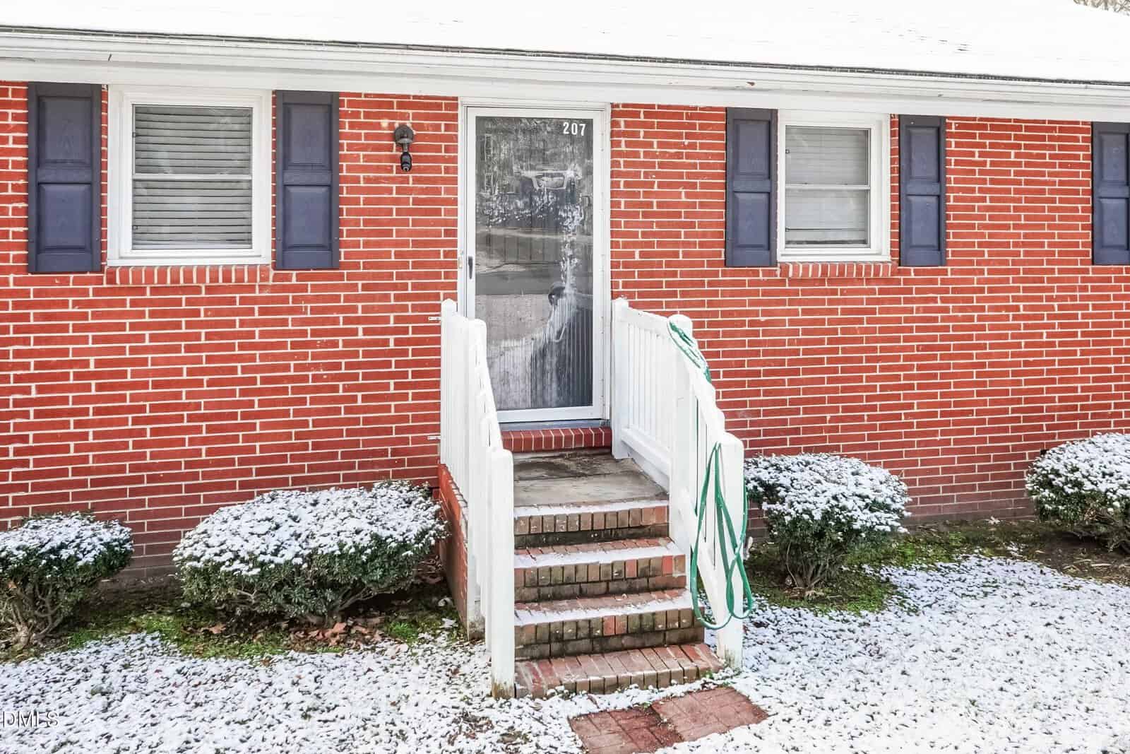 A front entrance to a red brick house with a white storm door, white railing, three steps, a green garden hose, two windows with blue shutters, and bushes dusted with snow.