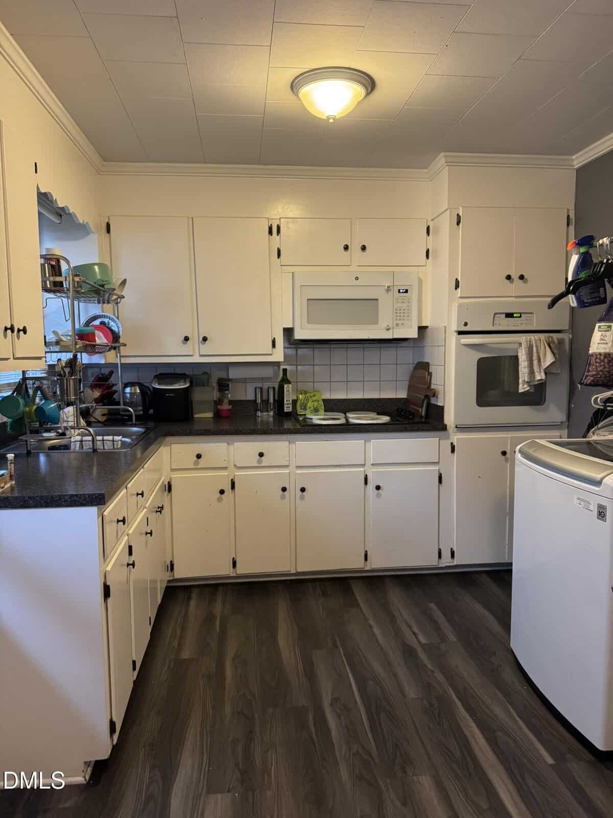 A kitchen with white cabinets, a microwave, an oven, black countertops, a dish rack, a coffee maker, and a standalone white freezer on a wood-patterned floor under a ceiling light.