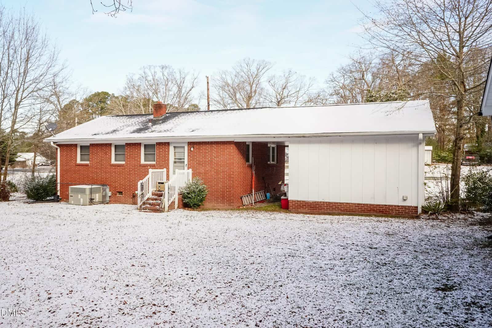 A red brick house with white trim and a small set of stairs leading to the back door stands in a yard dusted with a light layer of snow. The yard contains scattered shrubs and bare trees in the background.