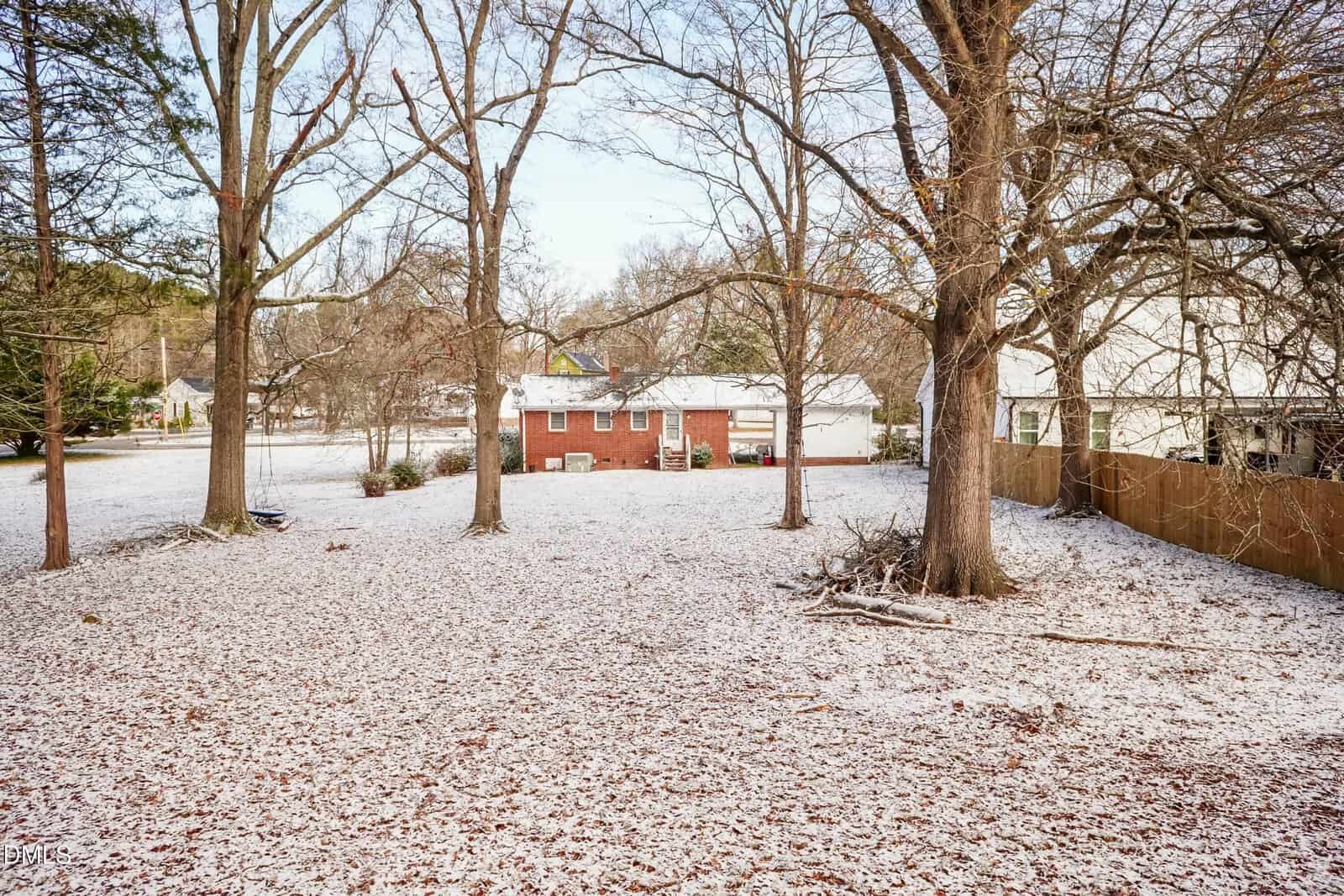 A backyard with large trees, light snow covering the ground, and a red brick house in the background. A wooden fence runs along the right side, and neighboring houses are visible beyond the yard.