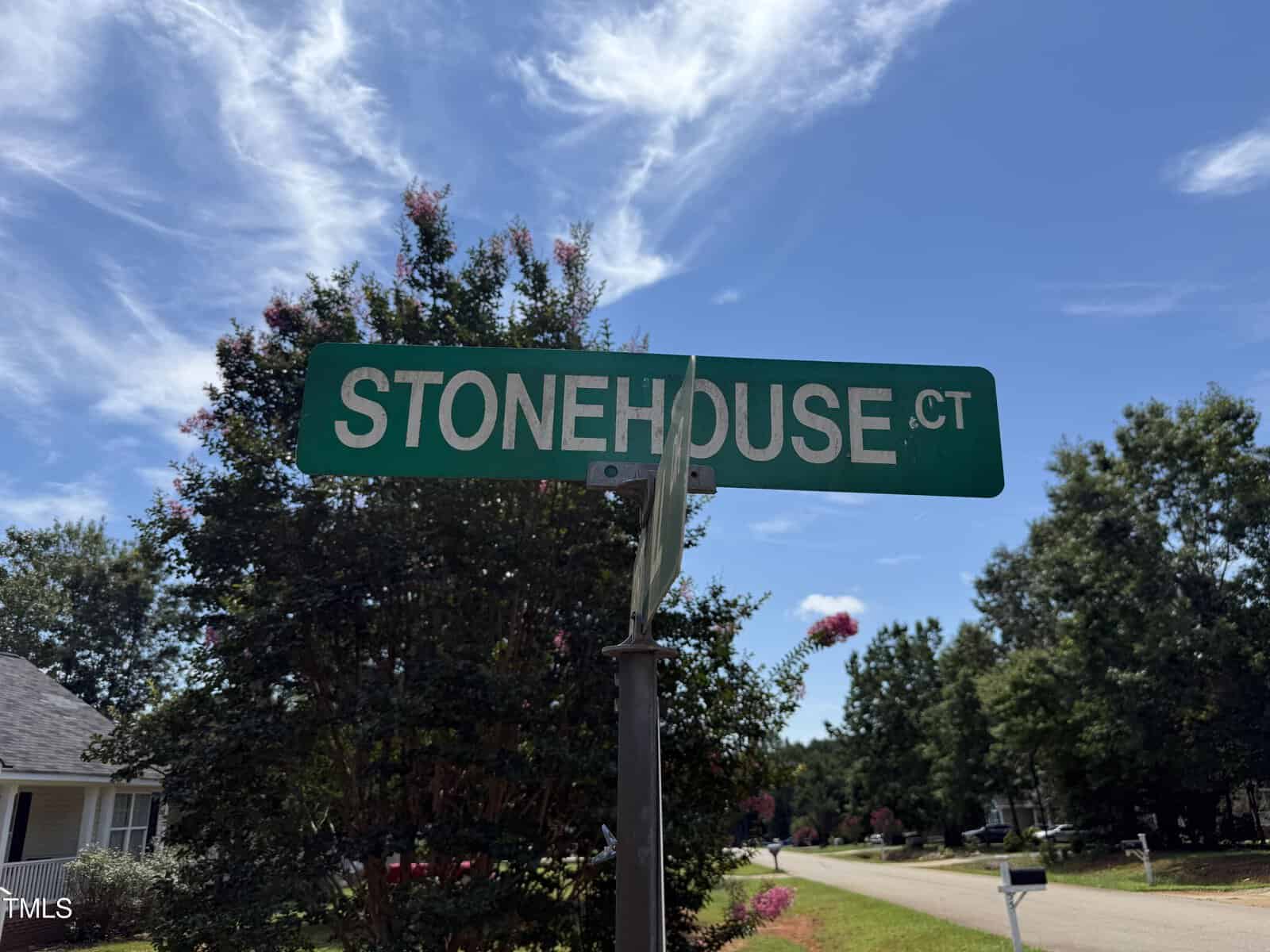 A green street sign reads "STONEHOUSE CT" against a blue sky, with trees and houses visible in the background on a sunny day.