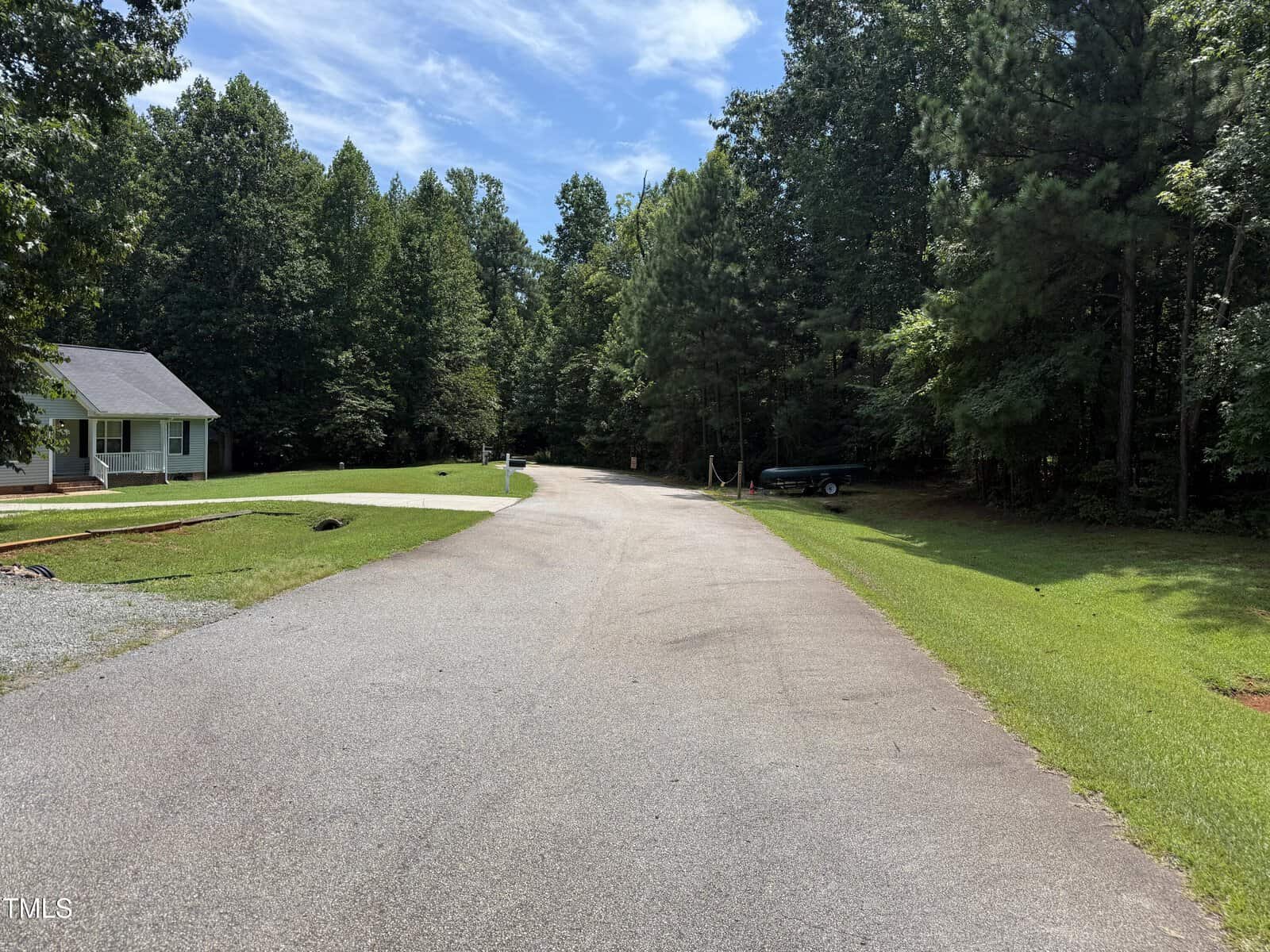 A quiet suburban street curves through a wooded area with green grass and tall trees; a small house with a porch sits on the left, and a parked boat is visible on the right. The sky is blue with some clouds.