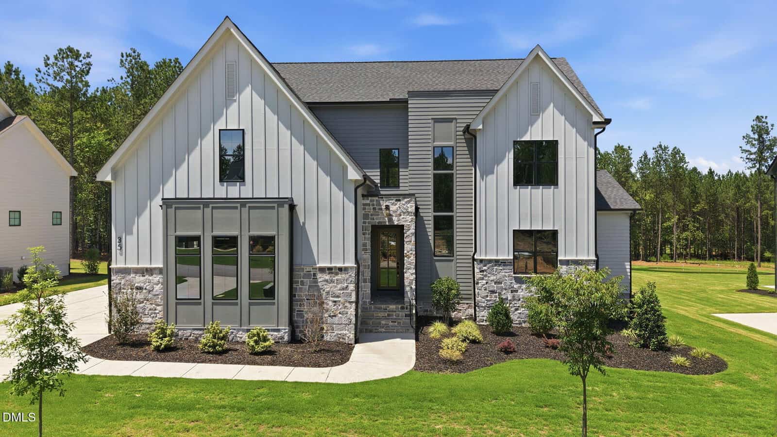 Modern two-story house at 35 Chestnut Oak Drive in Youngsville, featuring gray vertical siding, stone accents, large windows, and a front lawn with landscaped bushes and trees under a clear blue sky.