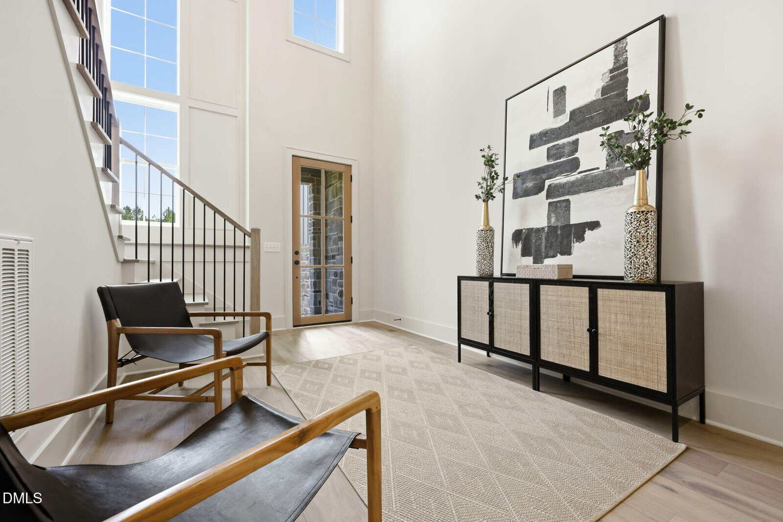 Modern, bright foyer with two black chairs, a patterned rug, a sideboard with wicker doors, two vases with green stems, and large abstract art. Staircase and tall windows bring in natural light.