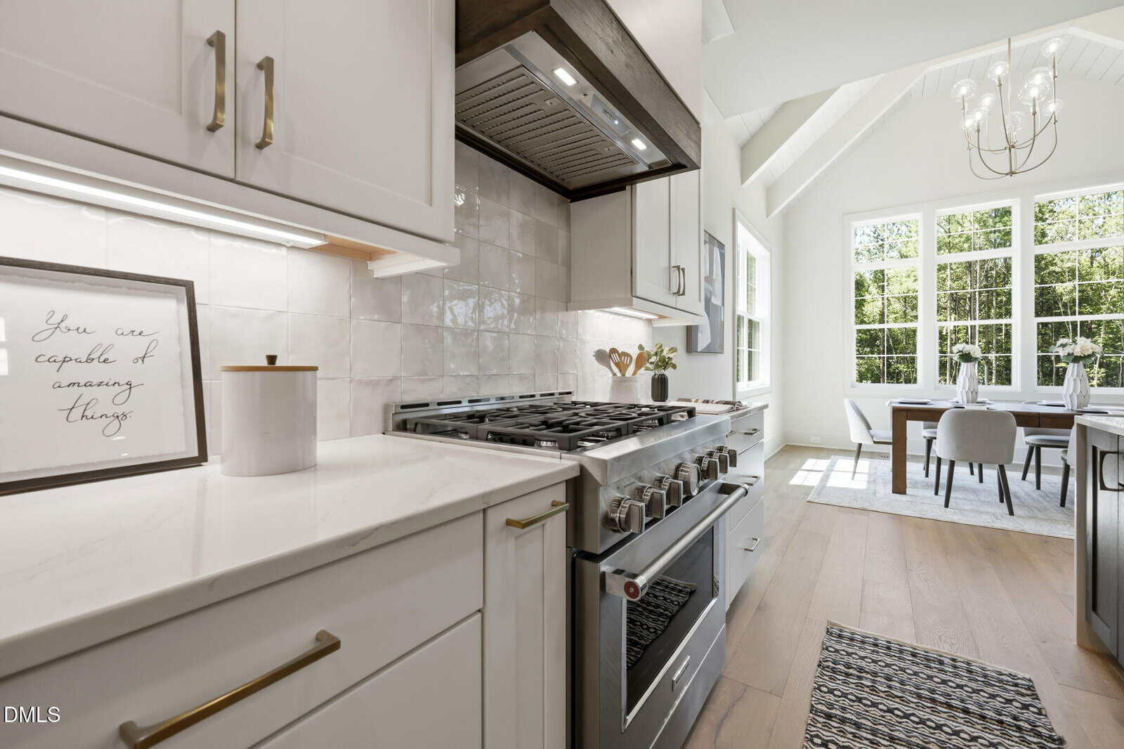 Modern kitchen with white cabinets, stainless steel stove, and a sign that says “You are capable of amazing things.” Bright dining area with large windows and a chandelier is visible in the background.